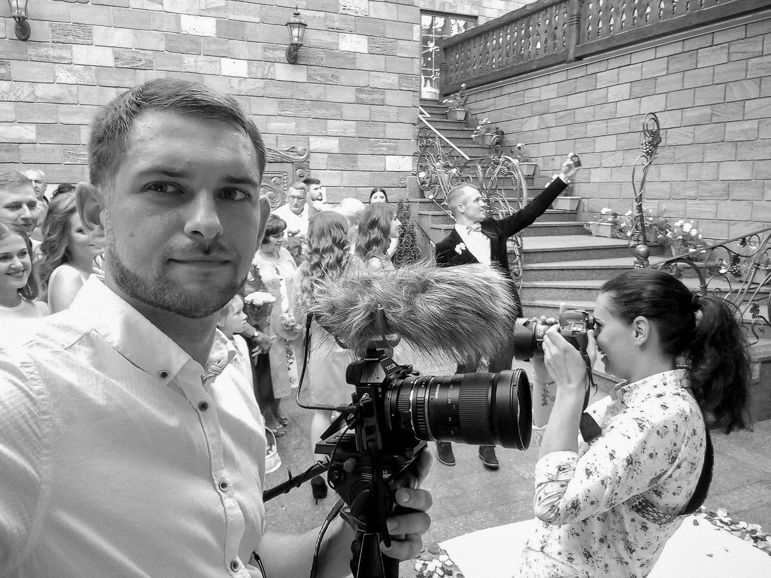 Black and white photo of a videographer holding a camera at an outdoor wedding ceremony with guests and couple on stairs.