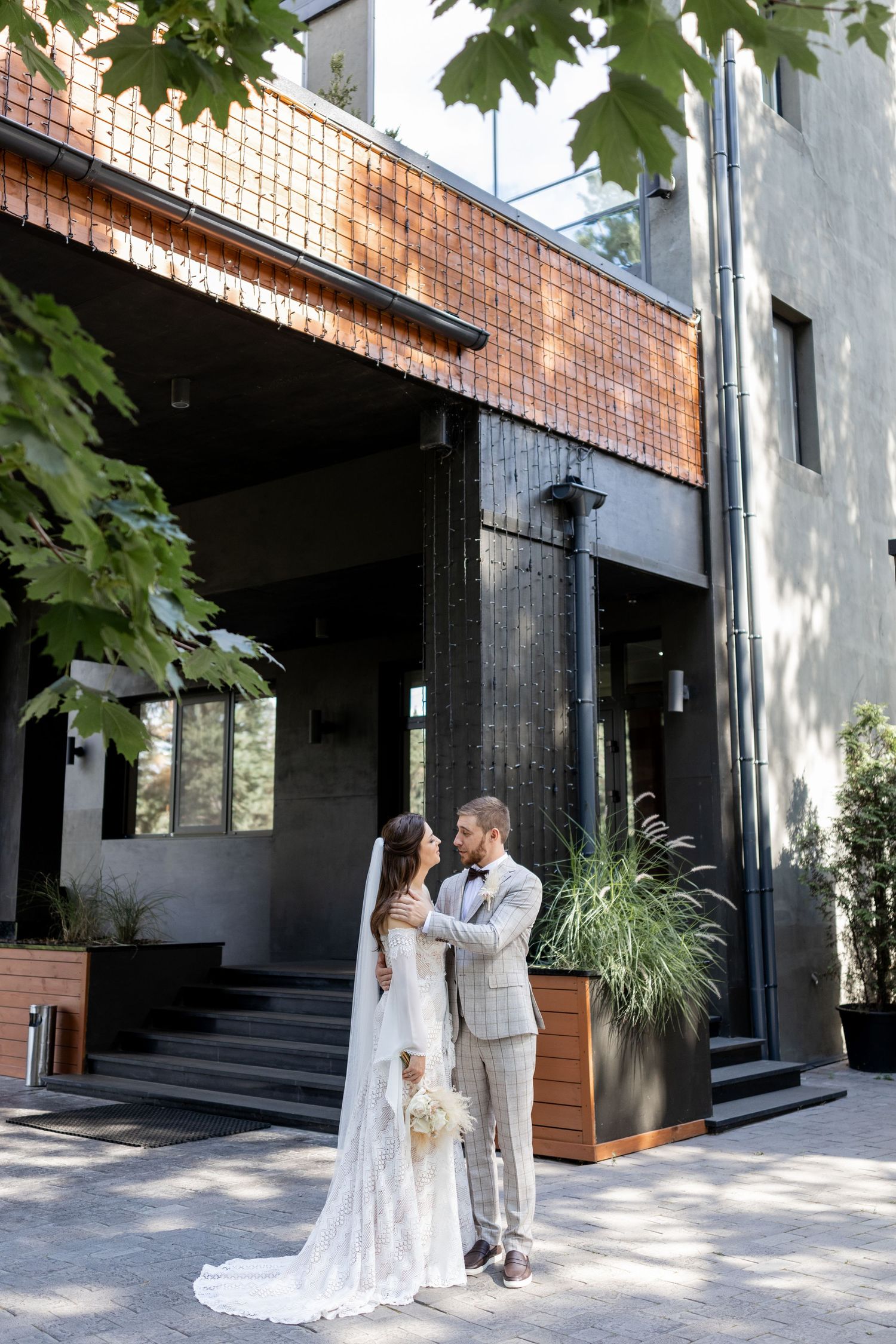 Wedding couple posing against modern brick and concrete architecture with maple leaves overhead.
