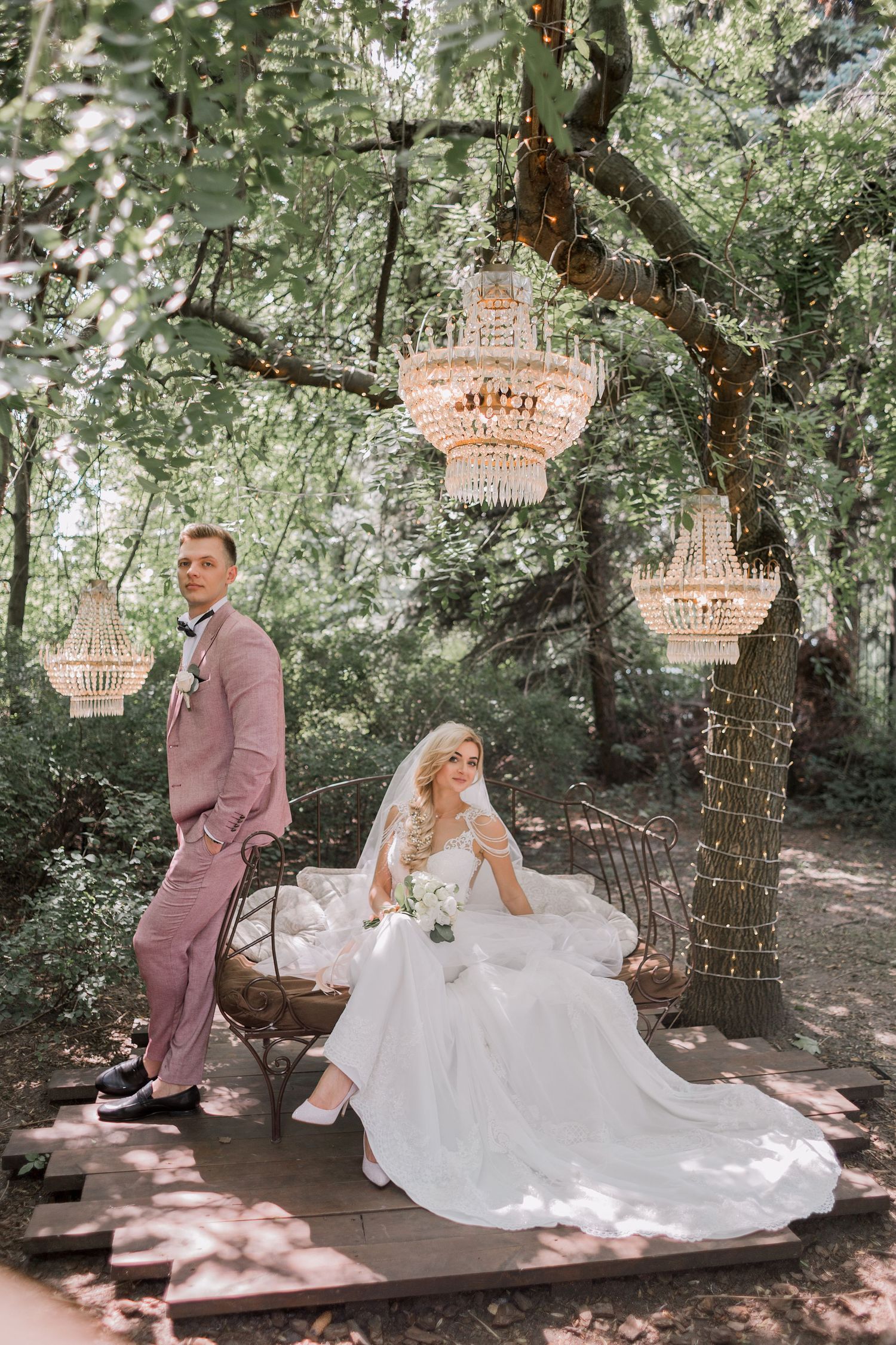 Bride in white gown sits on bench while groom in pink suit stands beside her under crystal chandeliers in lush garden.