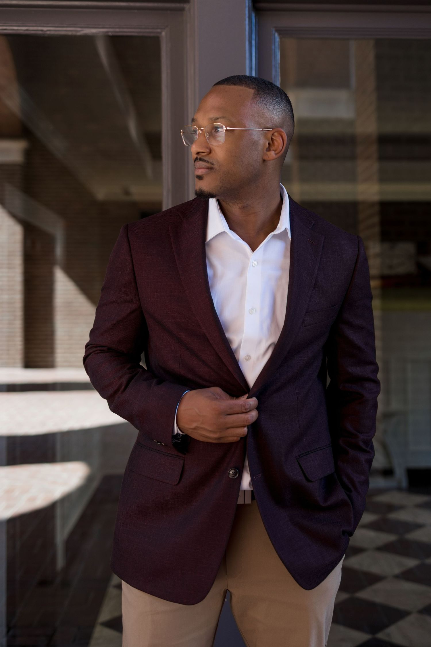 A stylish young Black man in a burgundy blazer and glasses poses confidently indoors.