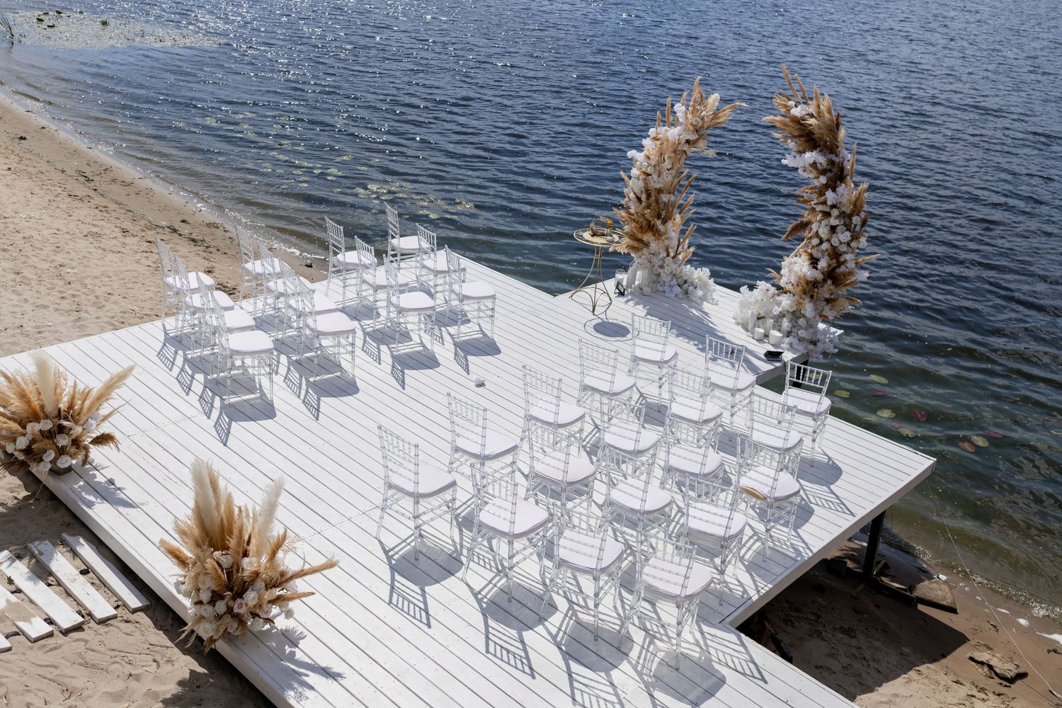 Elegant outdoor wedding ceremony setup on white dock by water with white chairs and floral arch.