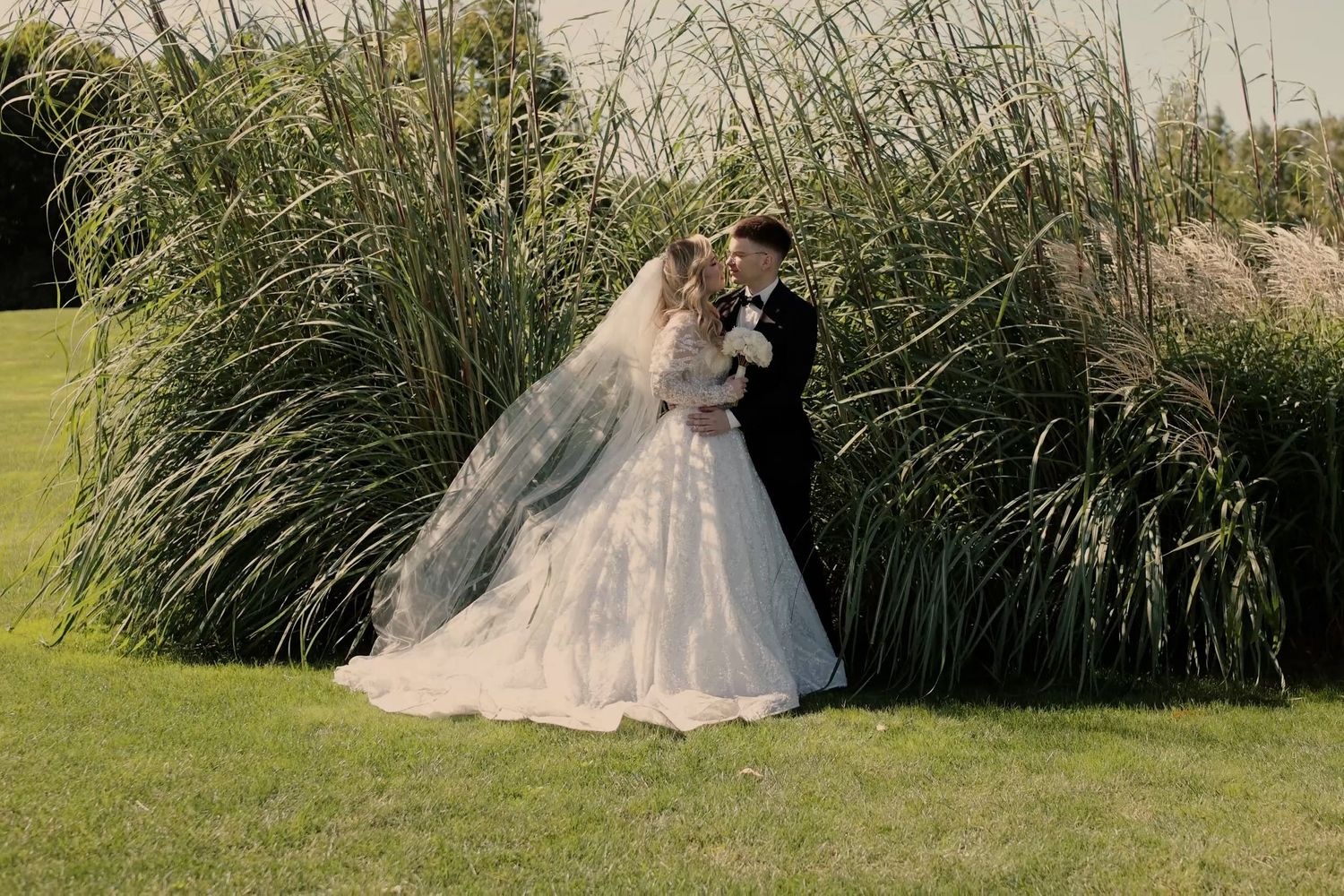 Bride and groom kissing in front of tall green grass, bride wearing a flowing white gown with veil.