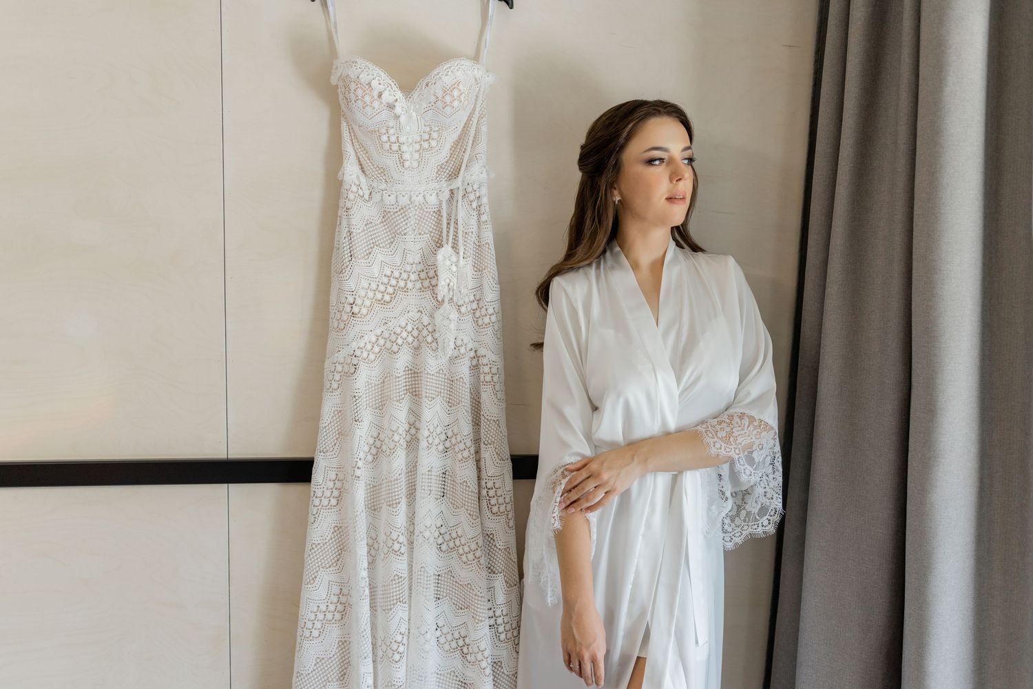 Wedding preparation scene showing a white lace dress hanging beside someone in a bridal robe.