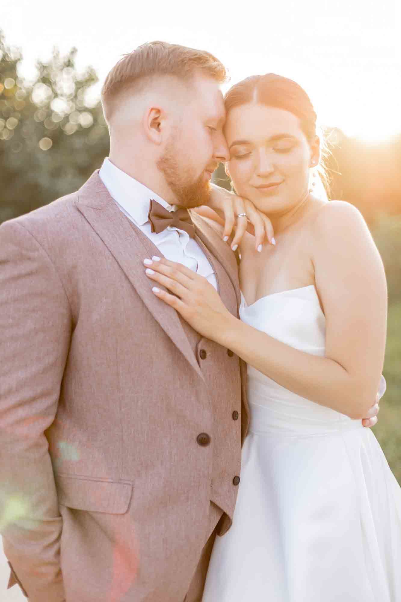 Bride and groom embrace during golden hour sunset, bride in strapless white gown, groom in pink suit with bow tie.