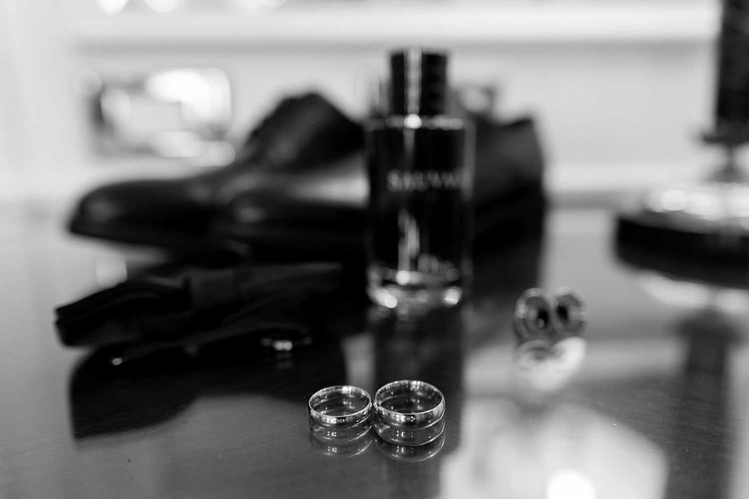 Black and white wedding flat lay with two rings, cologne bottle, dress shoes, and cufflinks on a reflective surface.