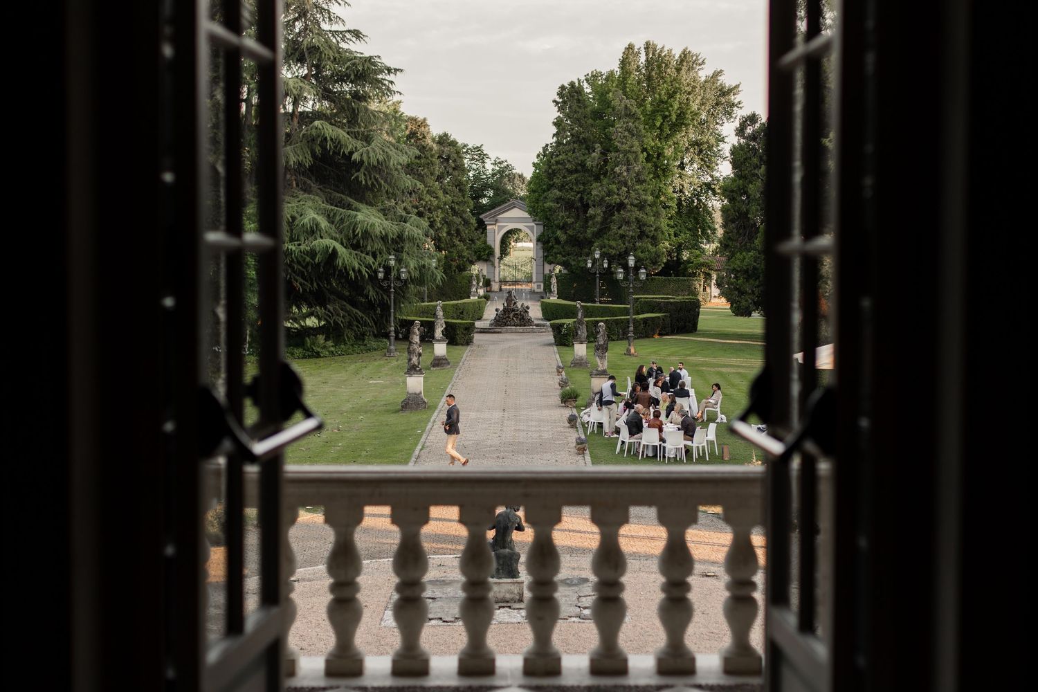 Elegant garden wedding venue viewed through ornate balcony doors, featuring manicured hedges and guests gathered near a classical gazebo.