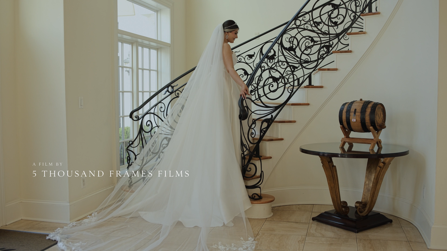 Bride in a flowing white gown and veil stands on an elegant staircase with ornate iron railings at a wedding venue.