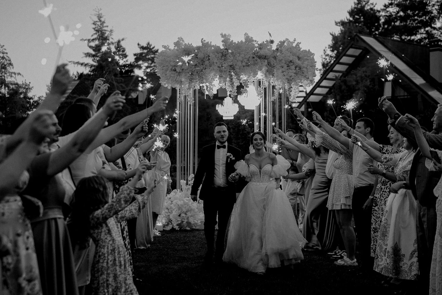 Black and white wedding photo of bride and groom walking through sparkler tunnel surrounded by cheering guests.