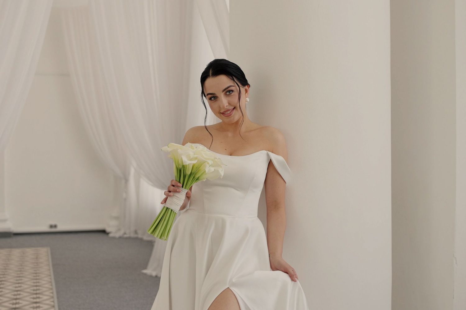 Elegant bride in white off-shoulder gown holding calla lily bouquet, posing gracefully between white columns.