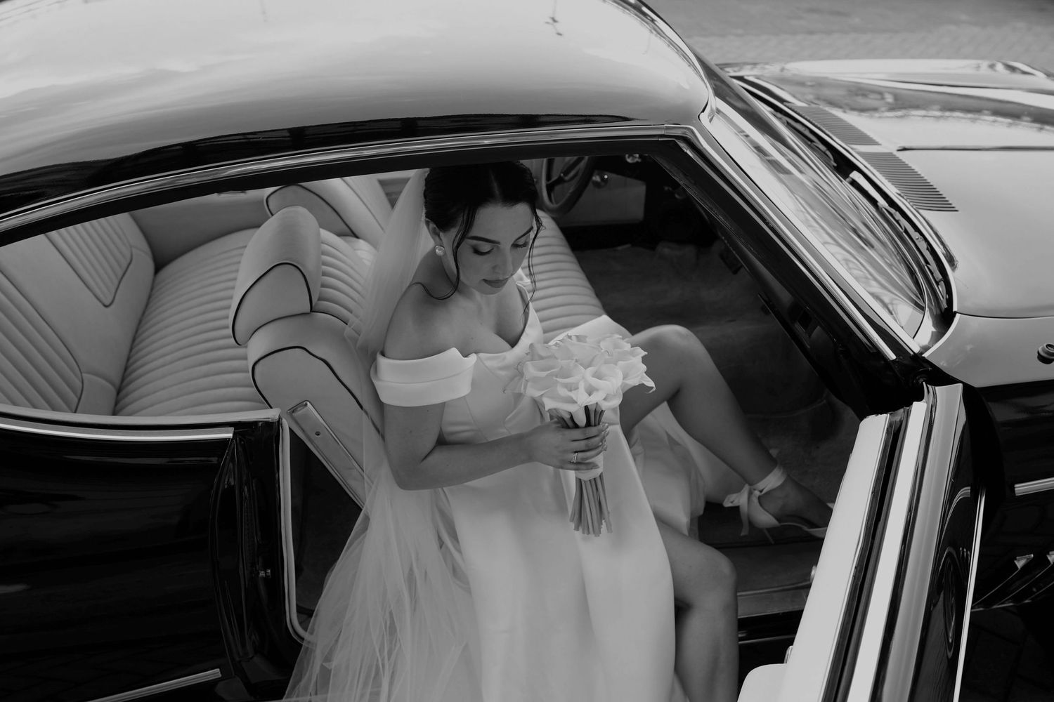 Bride in white off-shoulder gown holding bouquet, stepping out of vintage black car on wedding day.