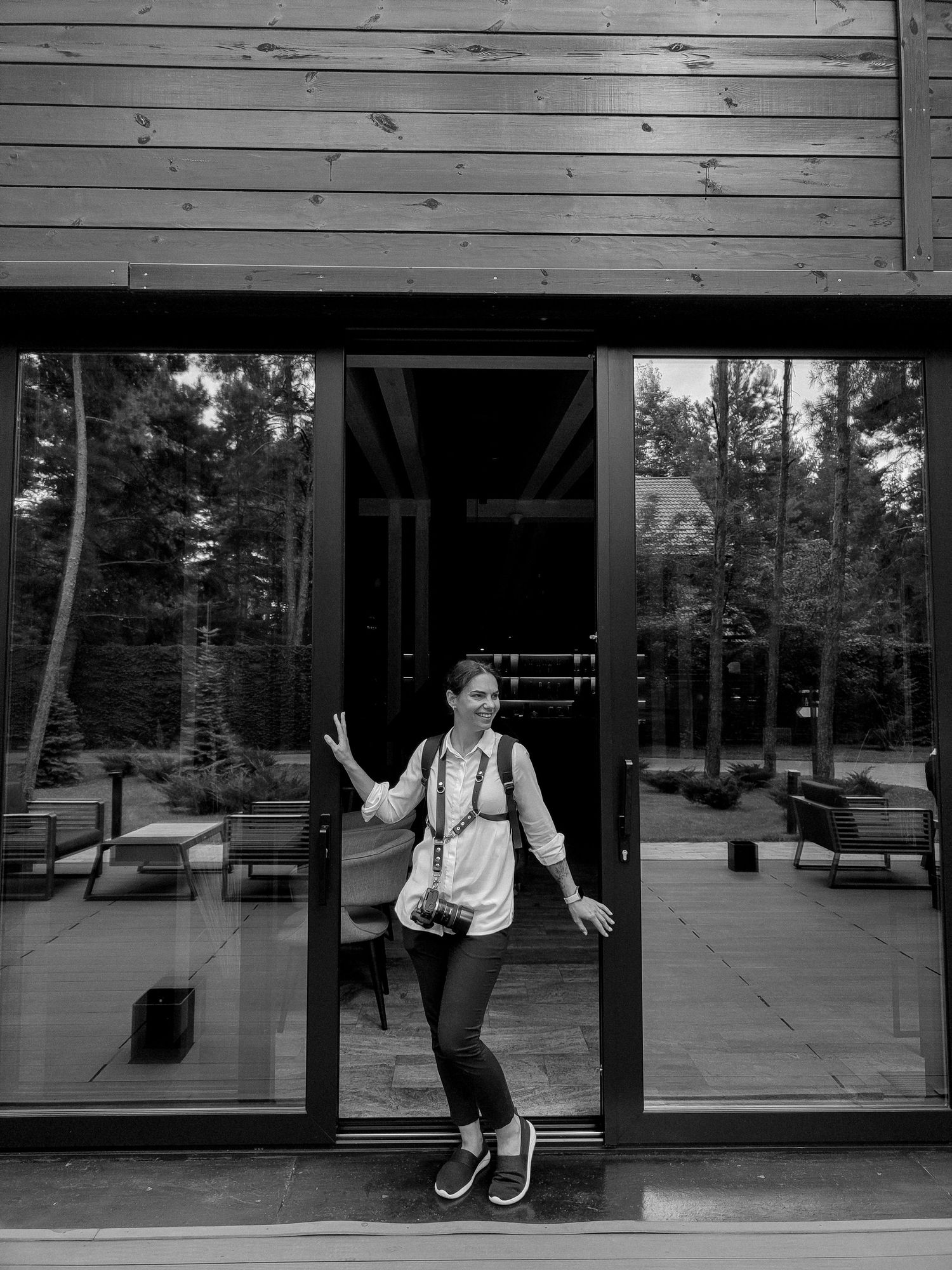 Black and white photo of a woman with a camera posing at glass doors of a modern wooden building surrounded by forest.