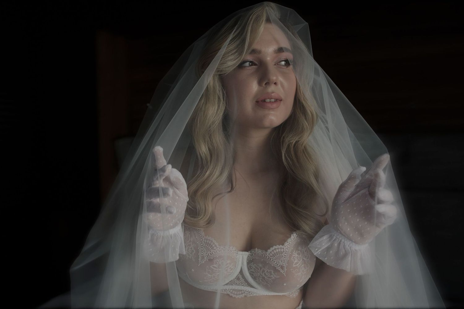 Woman in white bridal lingerie and veil with lace gloves posed dramatically in moody dark setting.