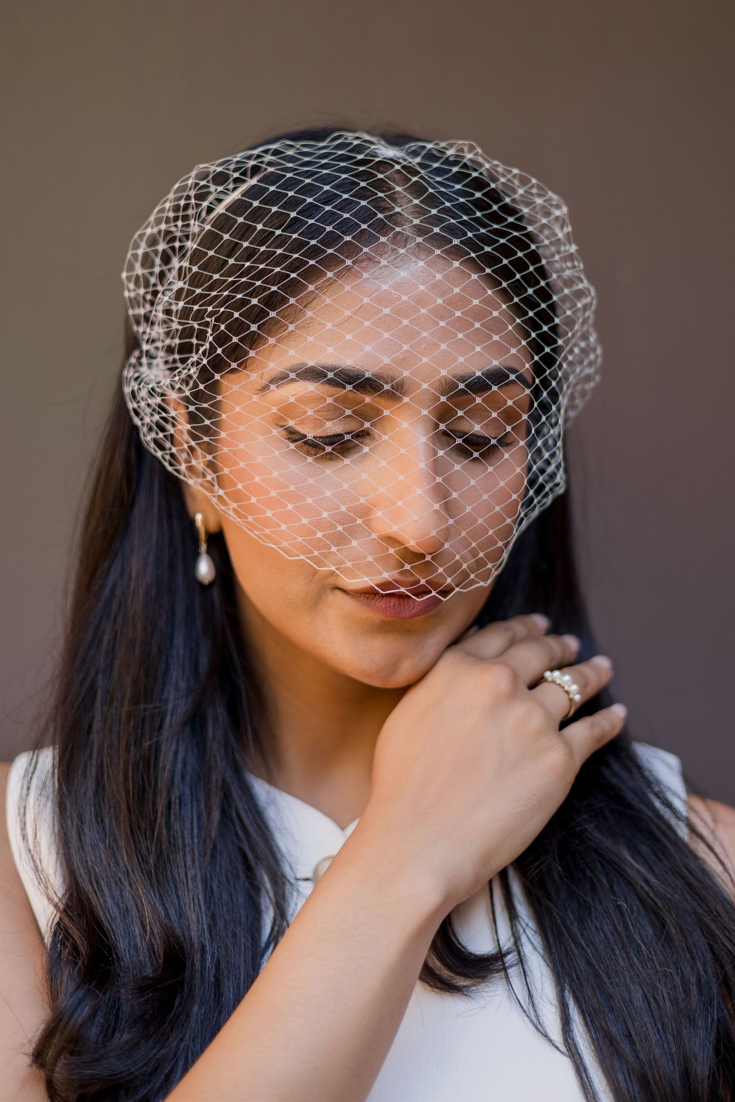 Bride wearing a delicate white birdcage veil with pearl earrings and ring, looking down gracefully.