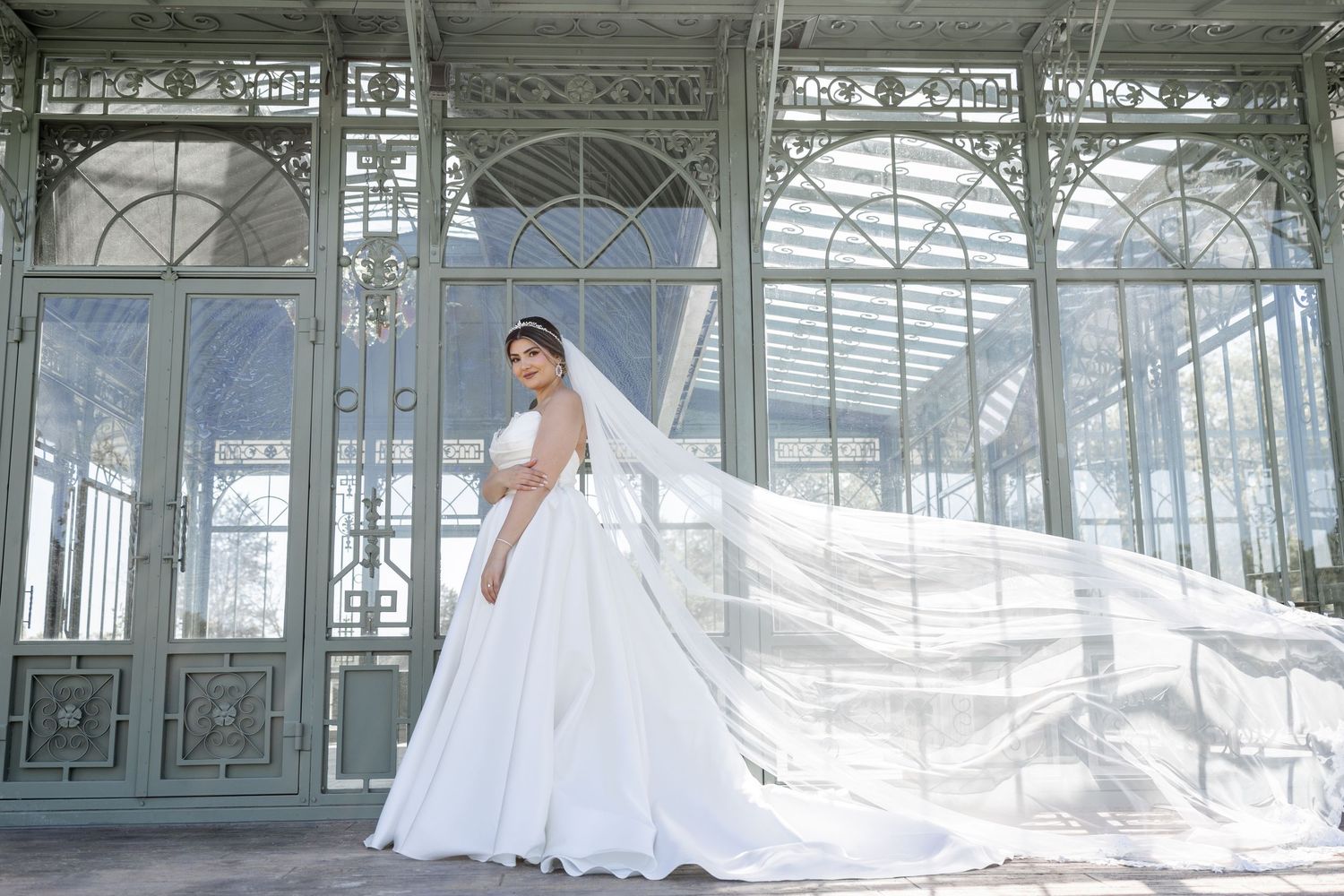 Bride in a flowing white ball gown with long veil in front of an elegant glass greenhouse.