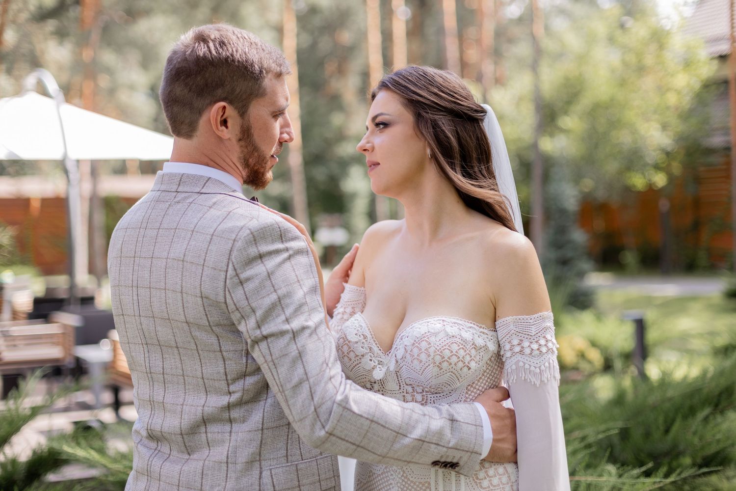 Romantic wedding couple embraces in garden setting with bride wearing off-shoulder lace dress.