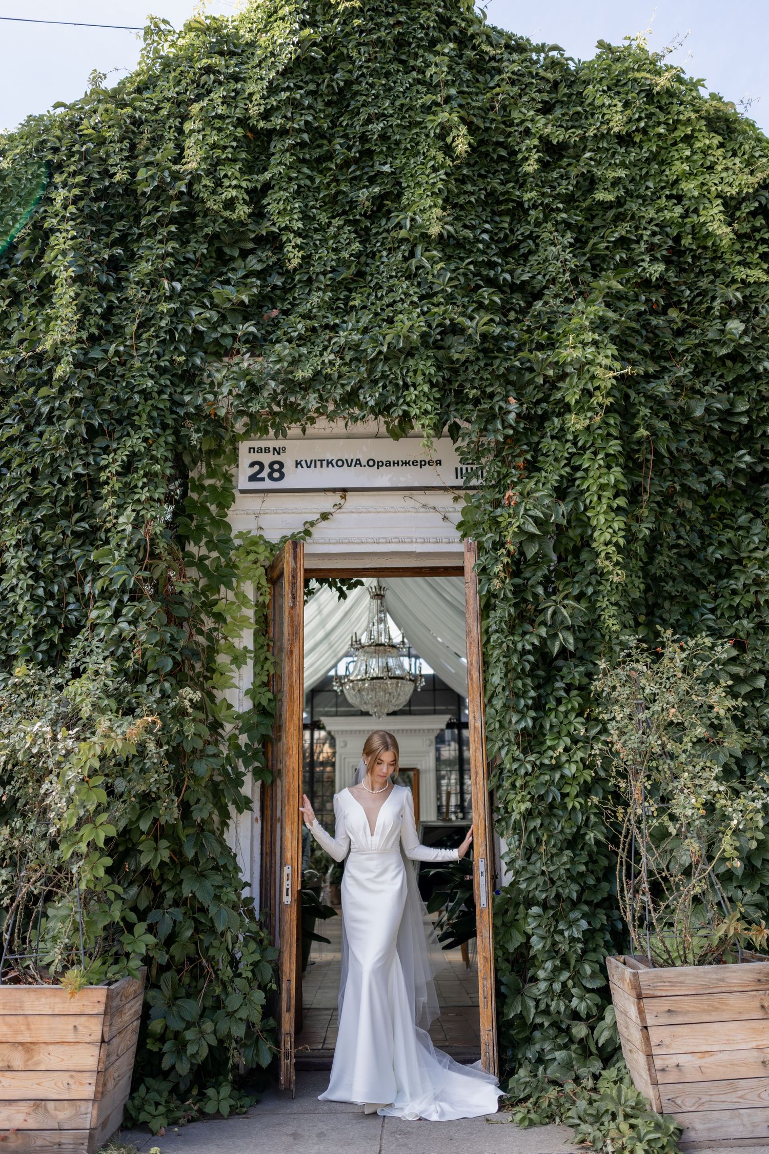 Elegant bride in long white gown standing in ivy-covered doorway at outdoor venue.