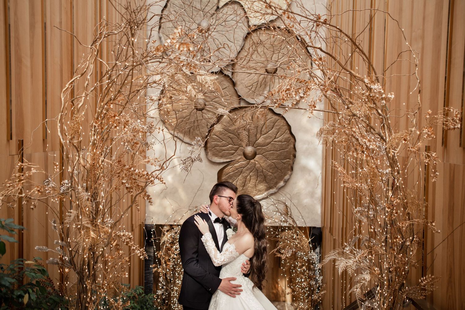 Bride and groom share a romantic kiss under a glittering branch arch with elegant floral backdrop.
