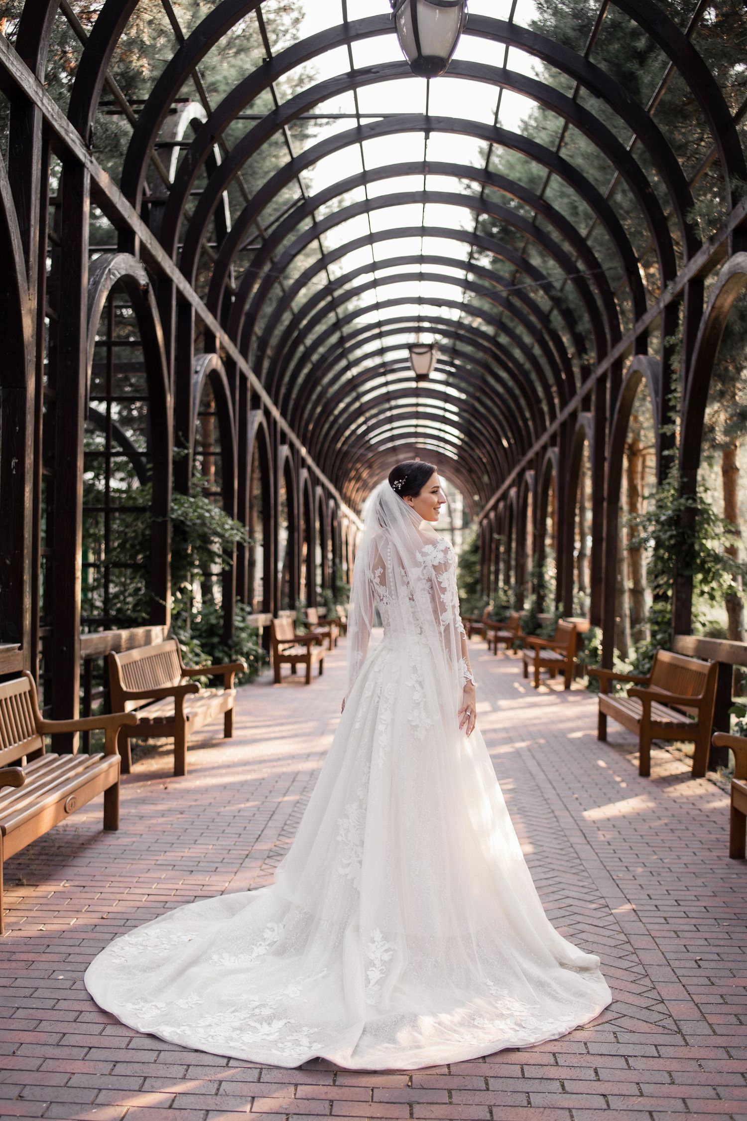 Bride in elegant lace wedding gown with veil stands in arched garden pergola with brick pathway.