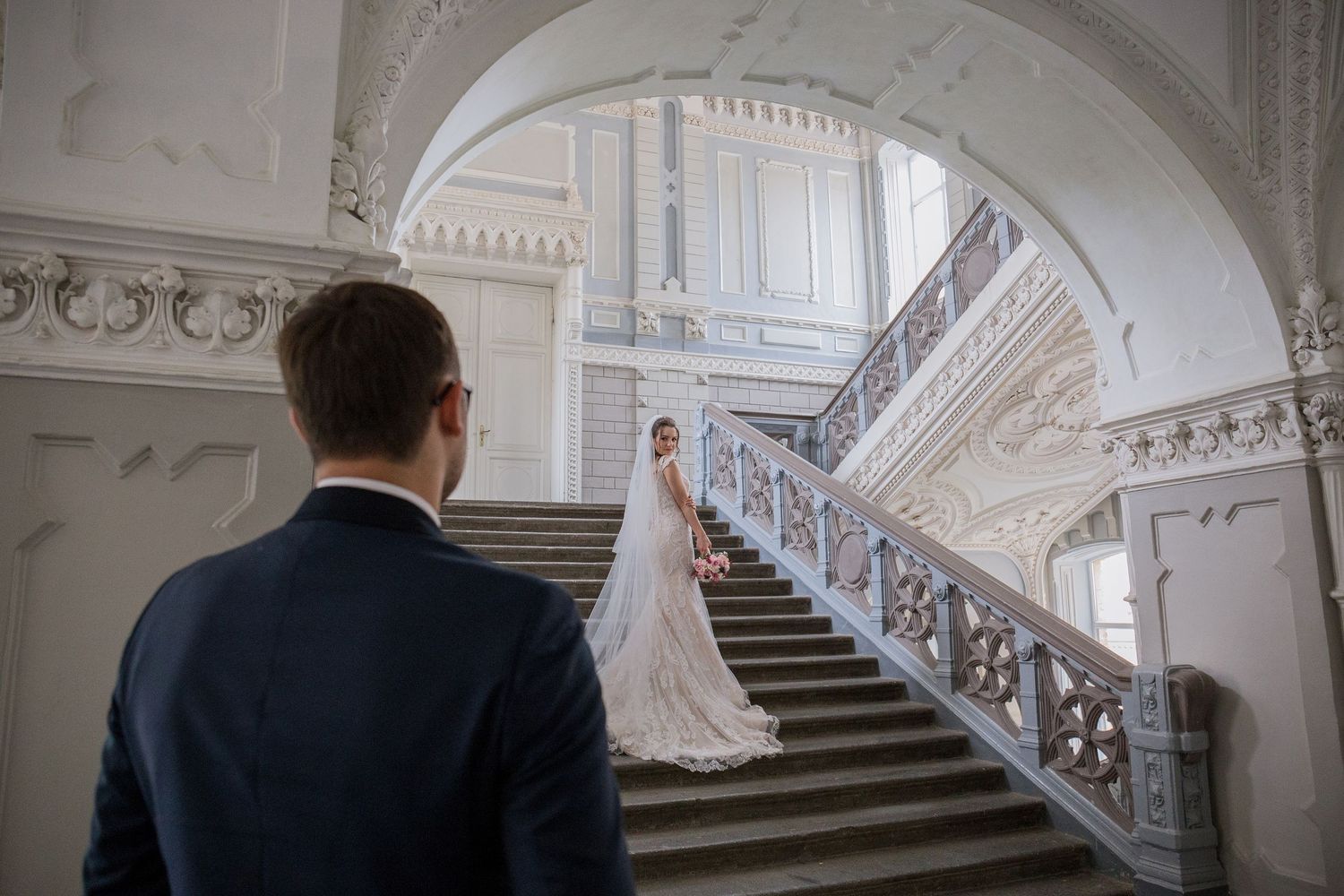 Bride in white gown stands on ornate staircase as groom in navy suit watches in elegant white wedding venue.