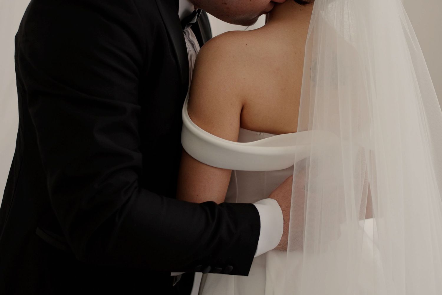 A groom in a black suit embraces a bride in a white off-shoulder gown and veil during their wedding.