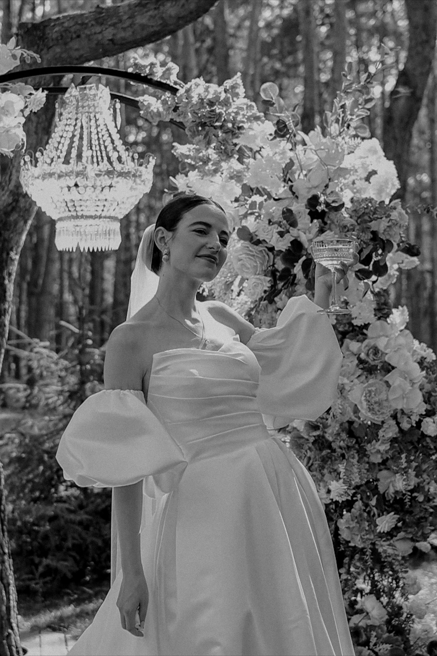 Bride in strapless white gown with puff sleeves poses by floral arch and chandelier in forest setting.