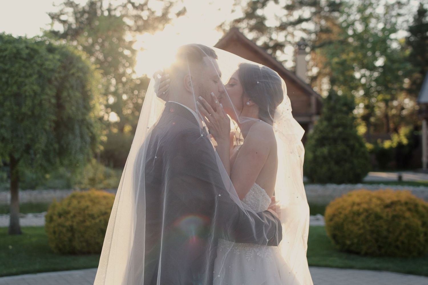 Bride and groom sharing a romantic kiss outdoors at golden hour, with soft sunlight glowing behind them at a garden wedding.