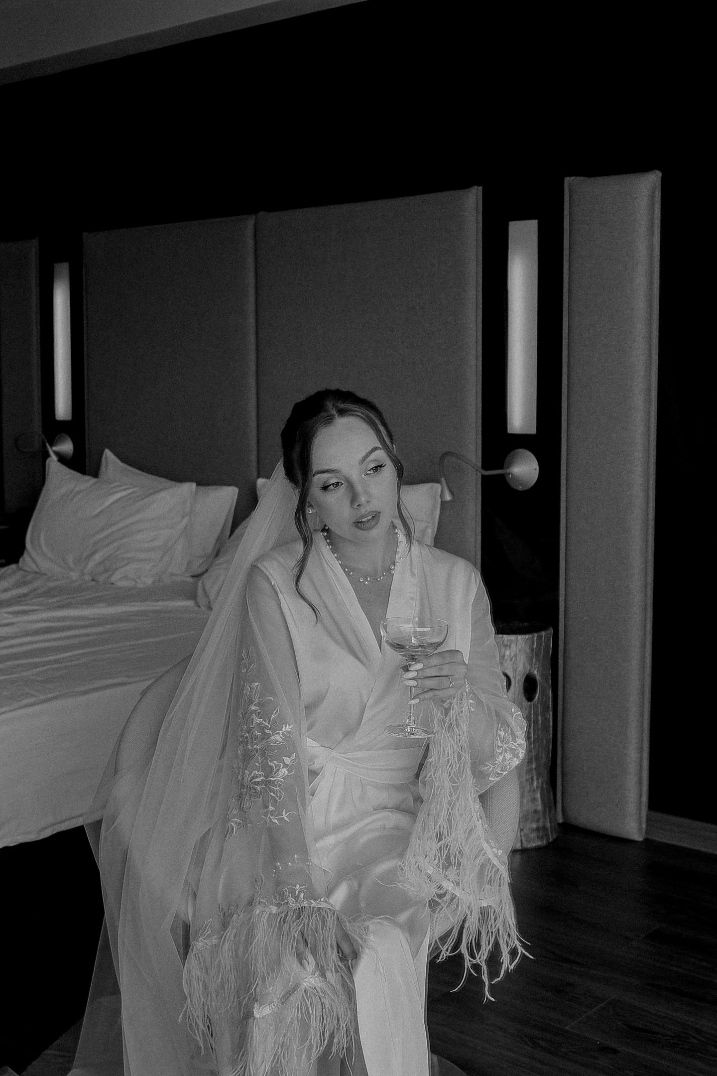 Black and white photo of a bride in a white feathered robe holding a wine glass, seated near a hotel bed.