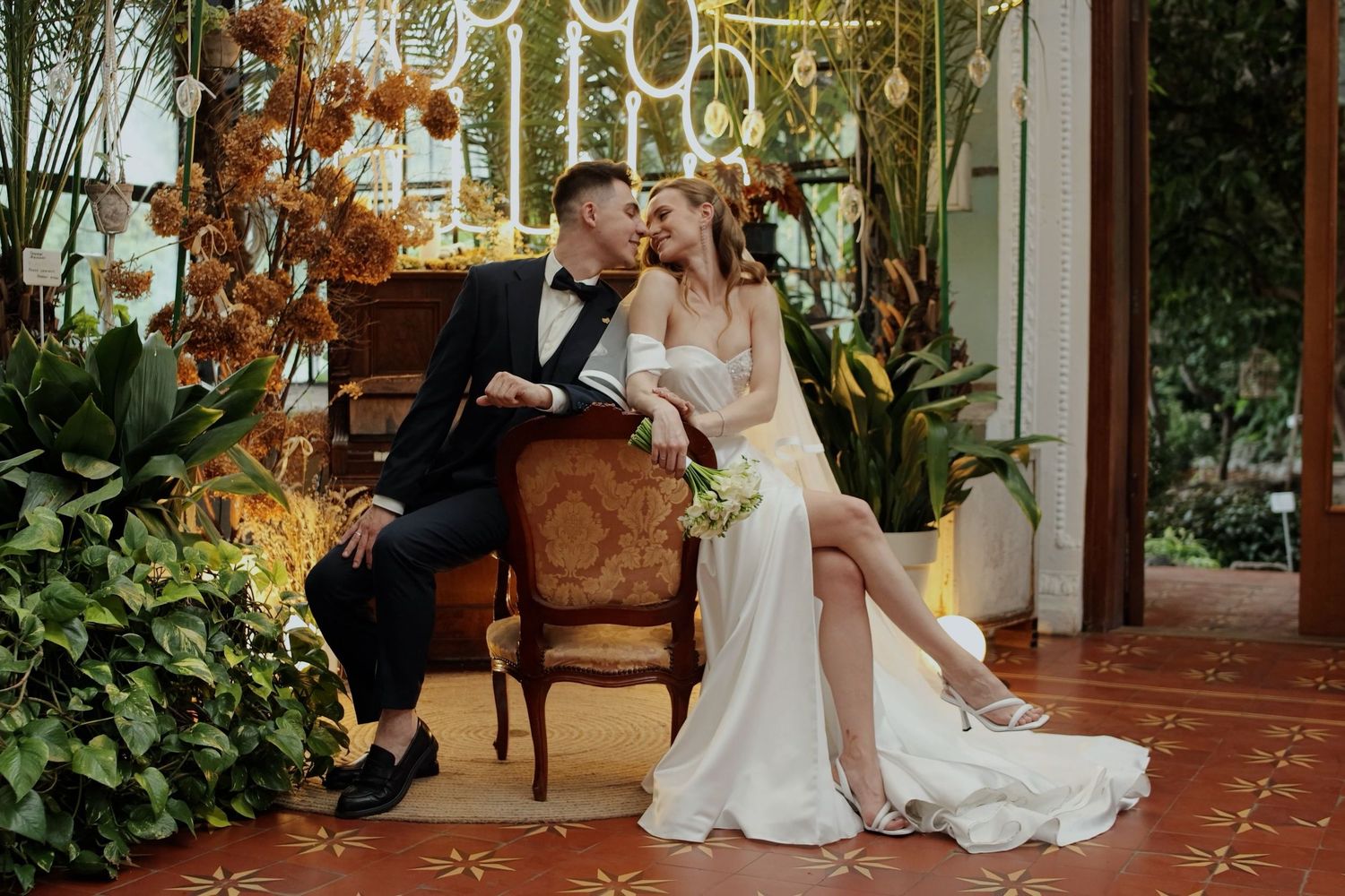 A bride and groom share a romantic moment in a lush botanical setting, with the bride in a white gown and groom in a tuxedo.