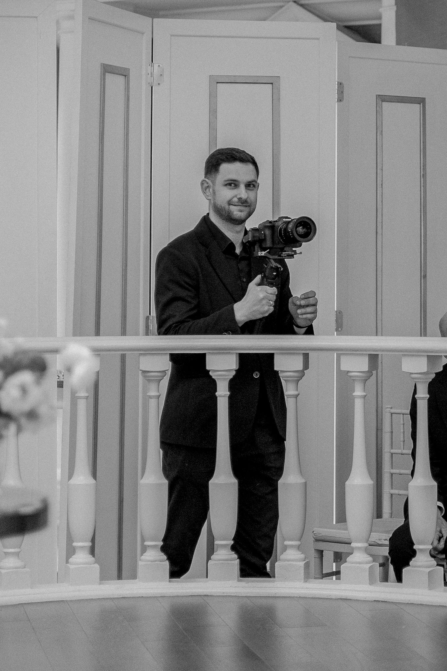 Black and white photo of a smiling man in a suit holding a professional camera on a stabilizer indoors.