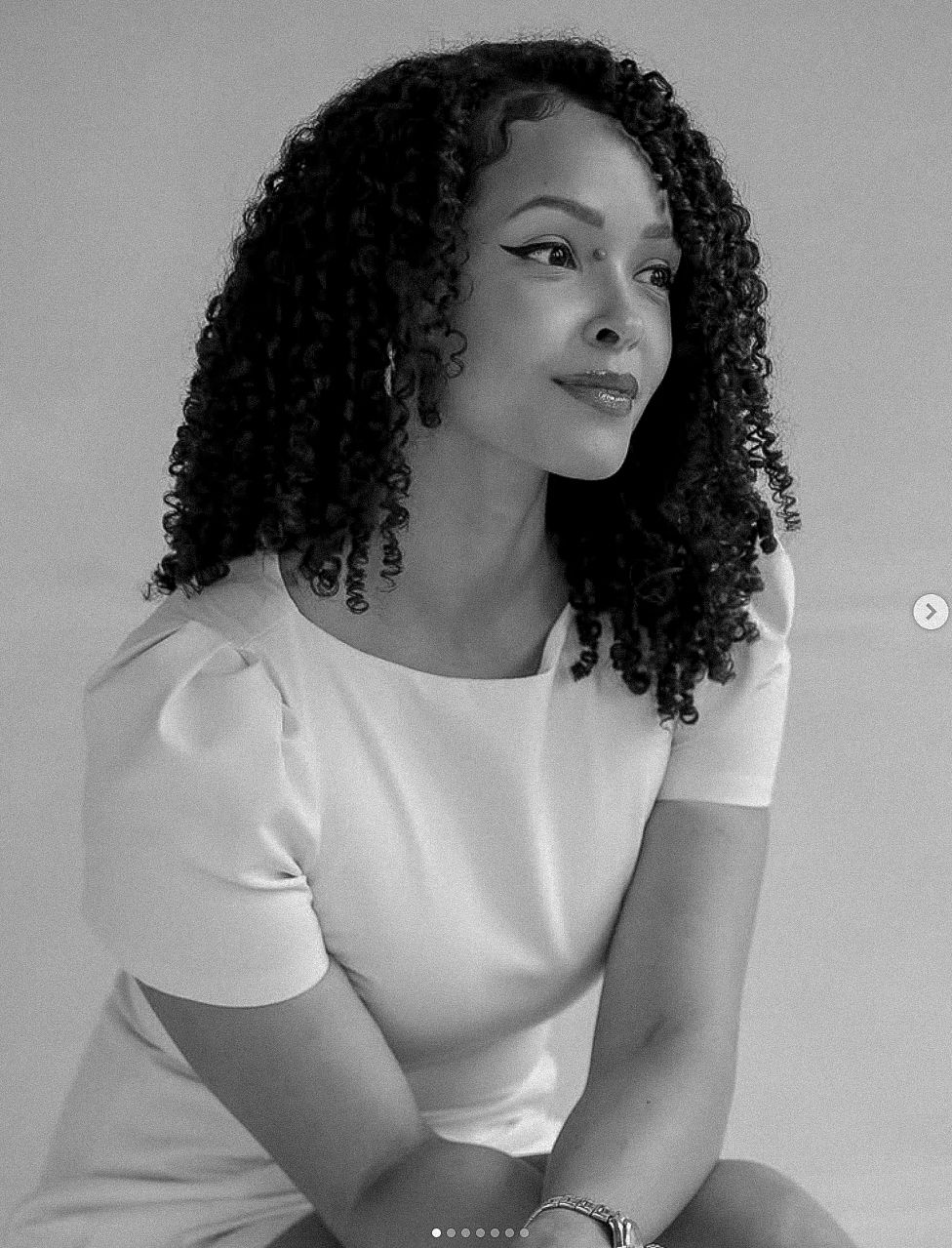 Black and white portrait of a young woman with curly hair wearing a white top, posed elegantly against a light background.
