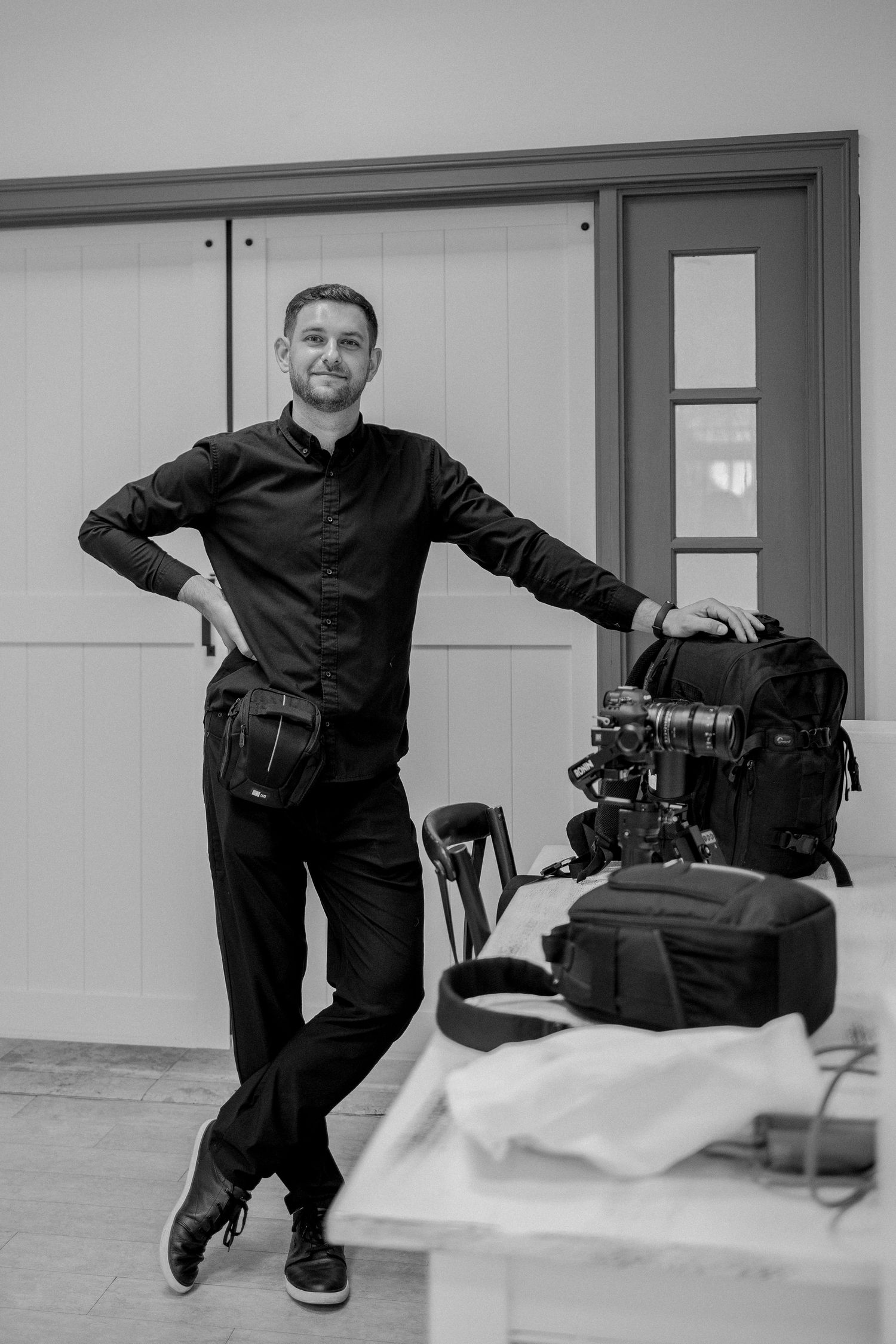 Black and white portrait of a smiling man in black attire posing with professional camera equipment indoors.