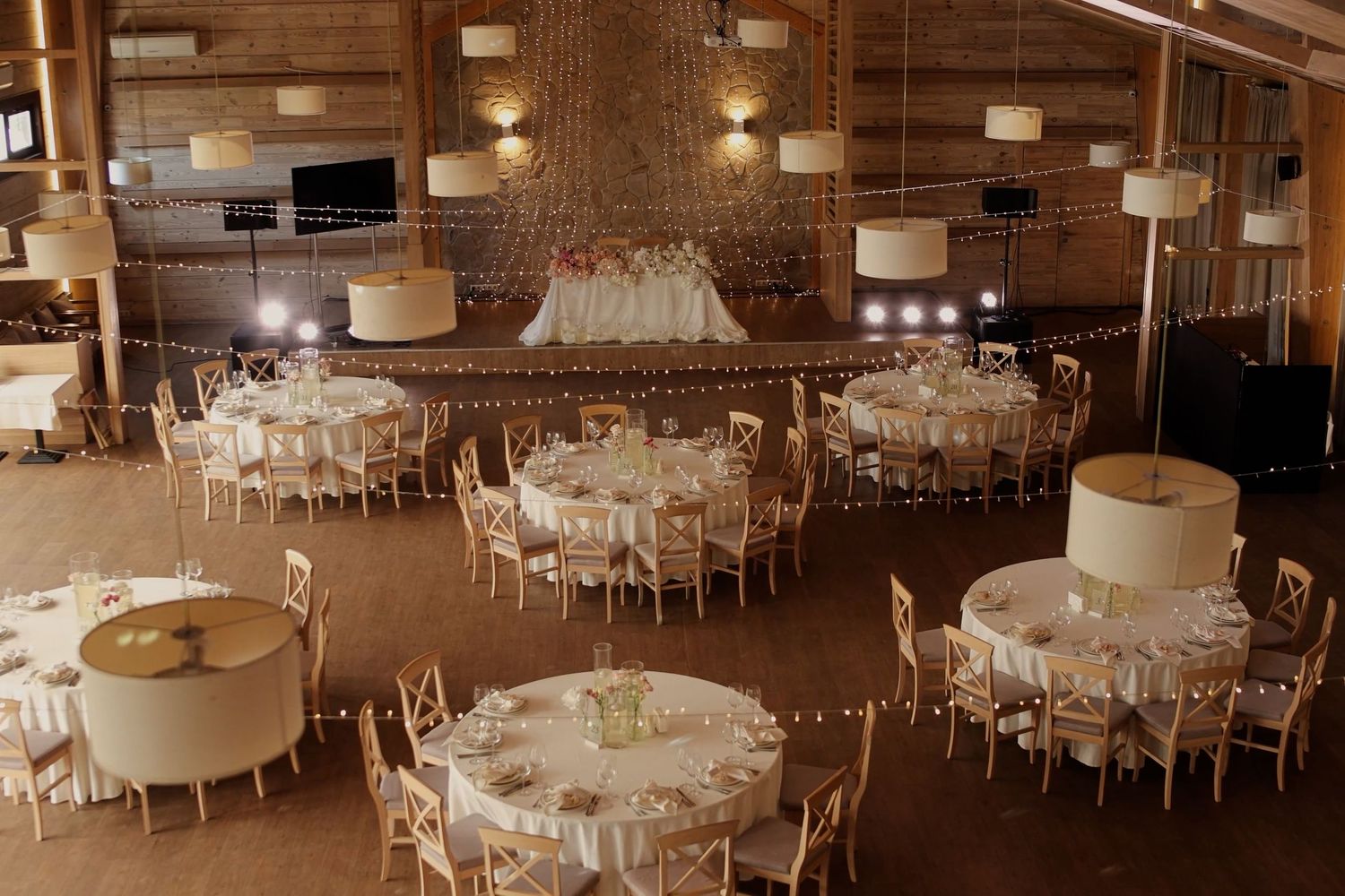 Elegant rustic wedding reception hall with round tables, white linens, string lights, and warm pendant lighting.