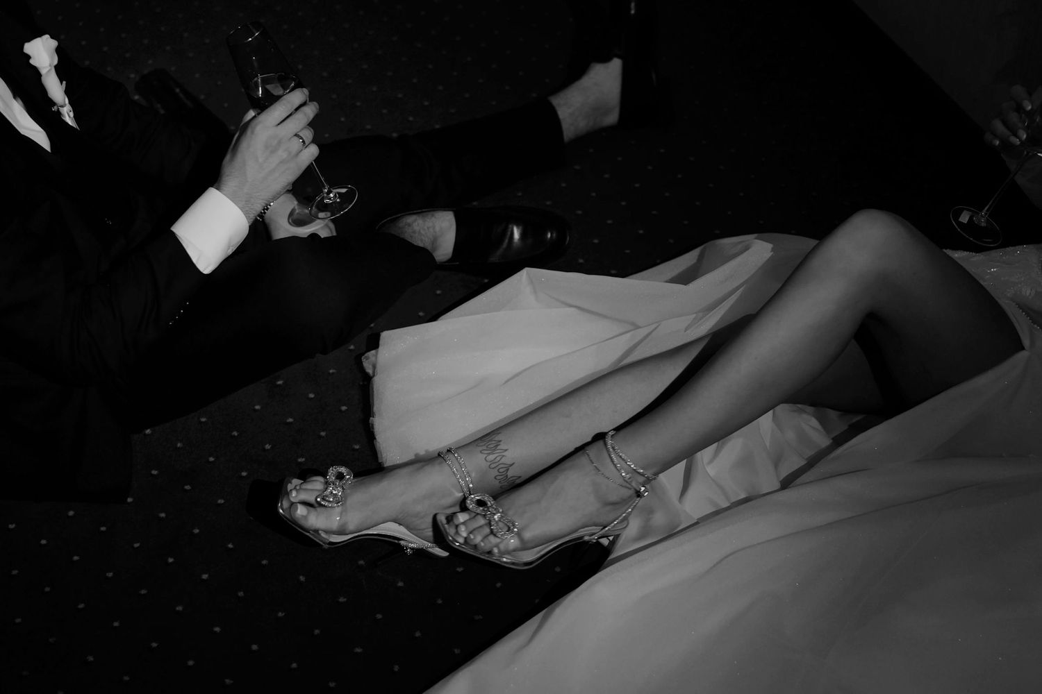Black and white photo of a bride in white gown with elegant heeled sandals, beside a suited guest holding a drink.