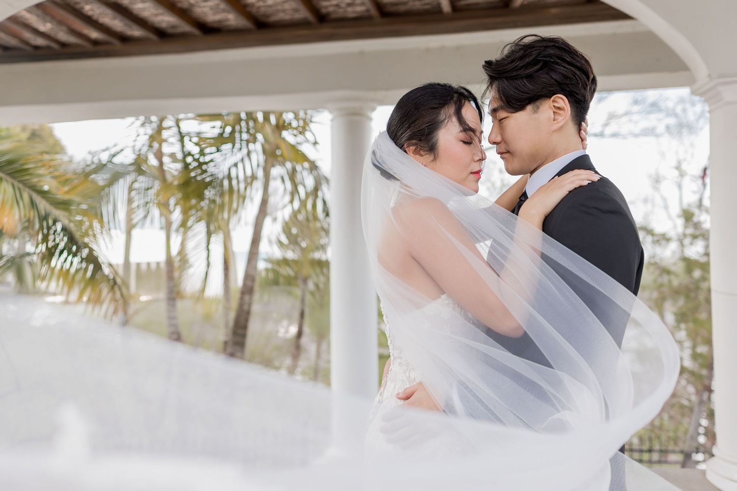 Asian couple in wedding attire share romantic moment under pavilion, bride's veil flowing in breeze with tropical backdrop.