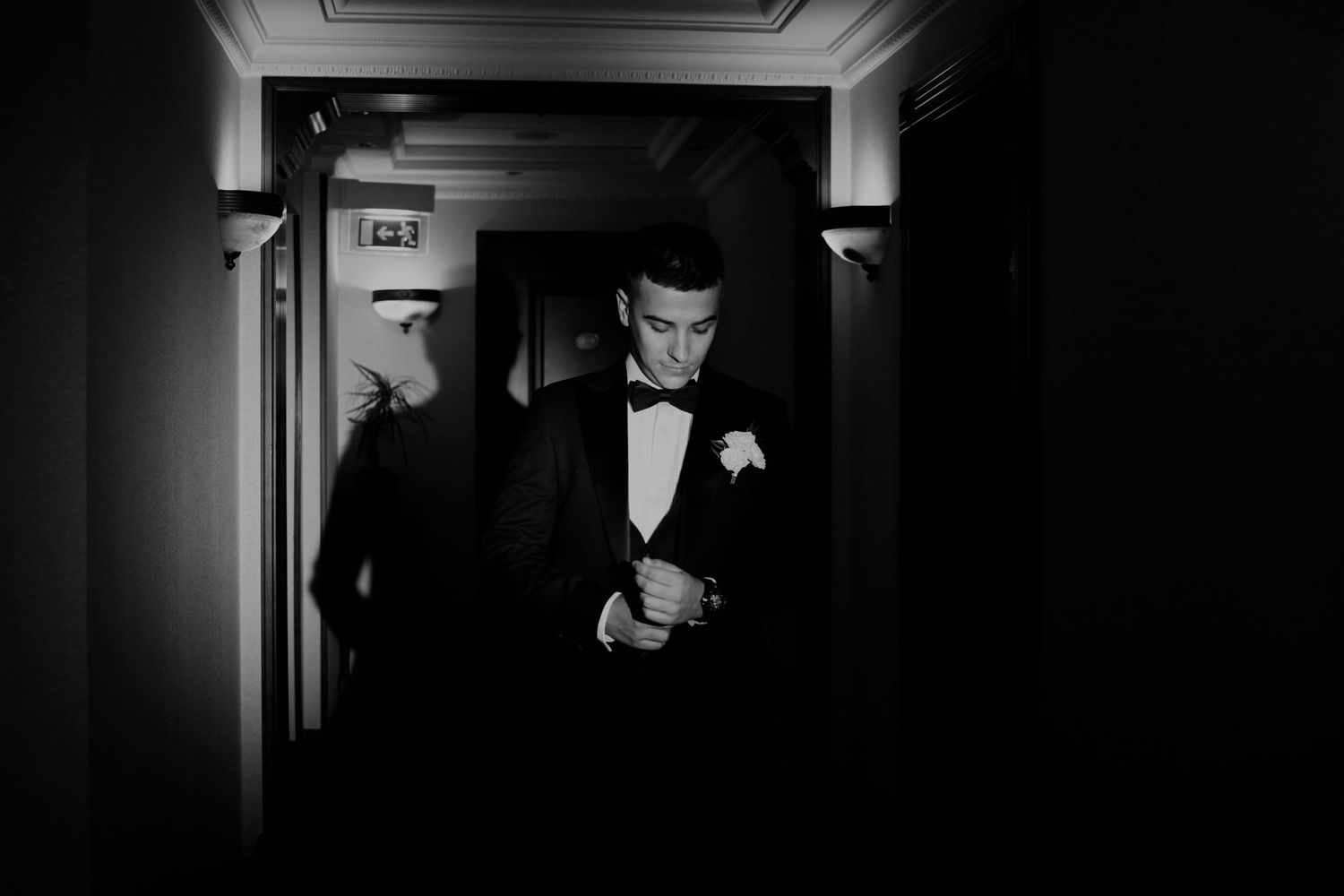 A groom in a black tuxedo adjusts his cufflinks in a dimly lit hallway in this elegant black and white wedding photo.