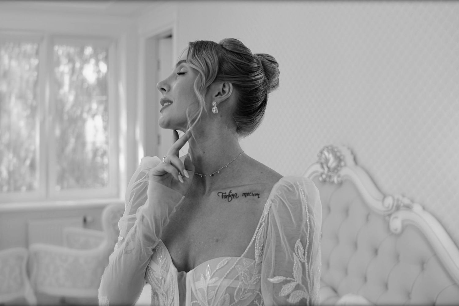 Black and white photo of a woman in a white lace dress with a neck tattoo, posing elegantly indoors.