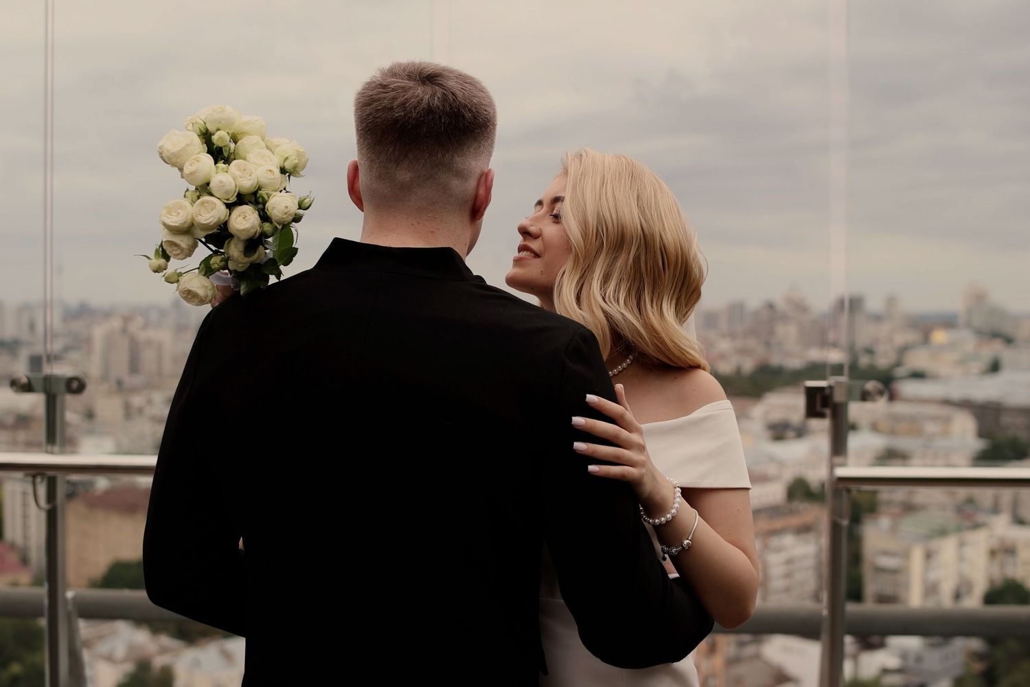 Couple embracing on rooftop with city skyline, groom holding white bouquet, bride in off-shoulder dress.