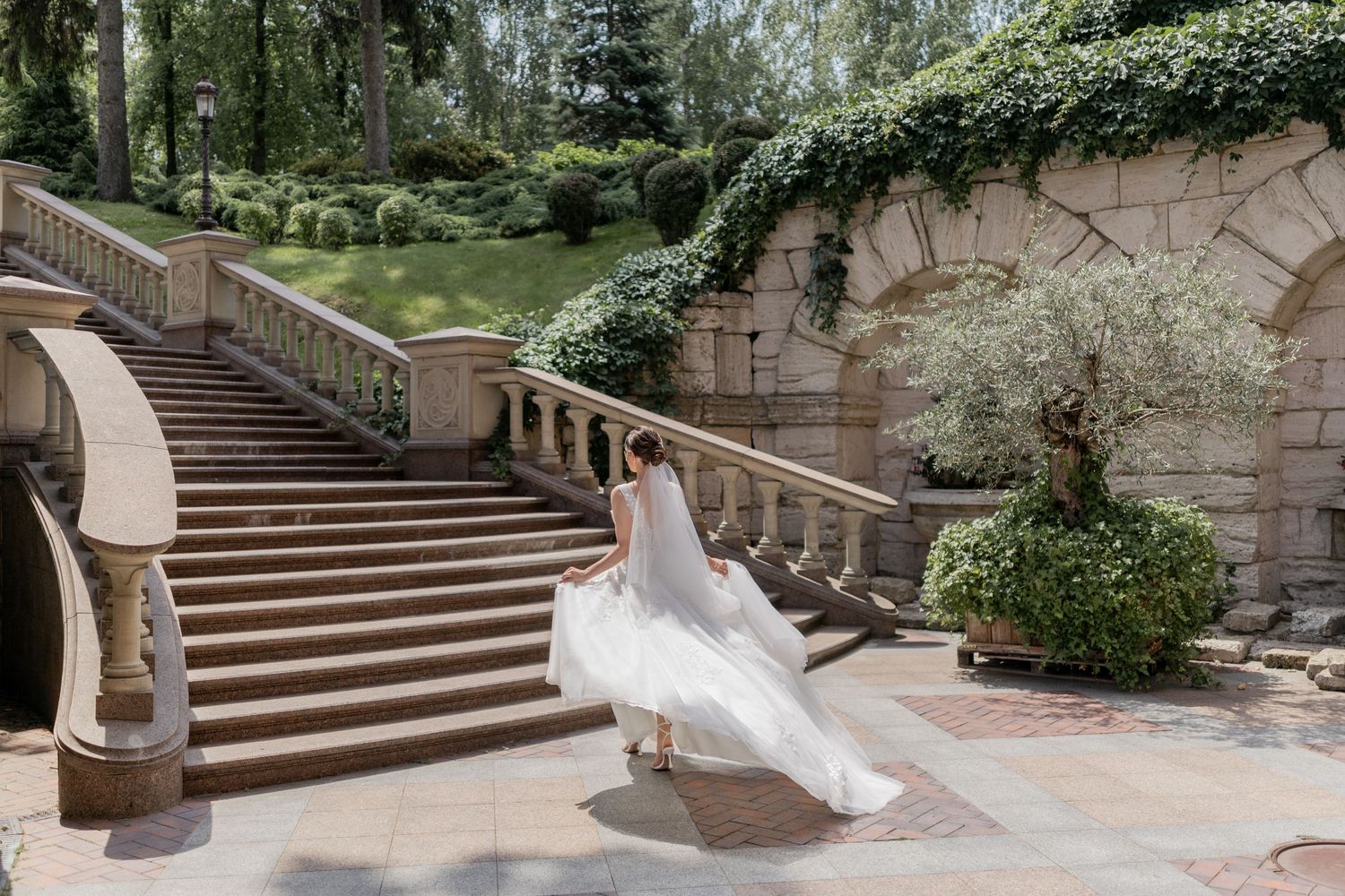 Bride in flowing white wedding gown twirling on stone patio beside elegant staircase surrounded by lush greenery.