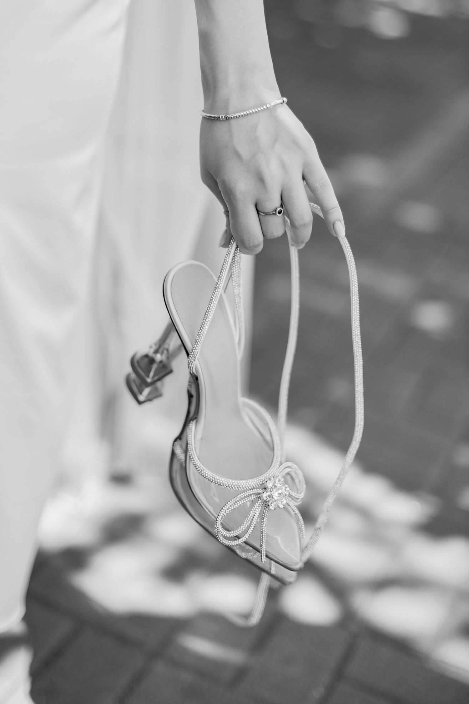 Black and white photo of a bride holding elegant strappy heels with bow detail, wearing a ring and bracelet.