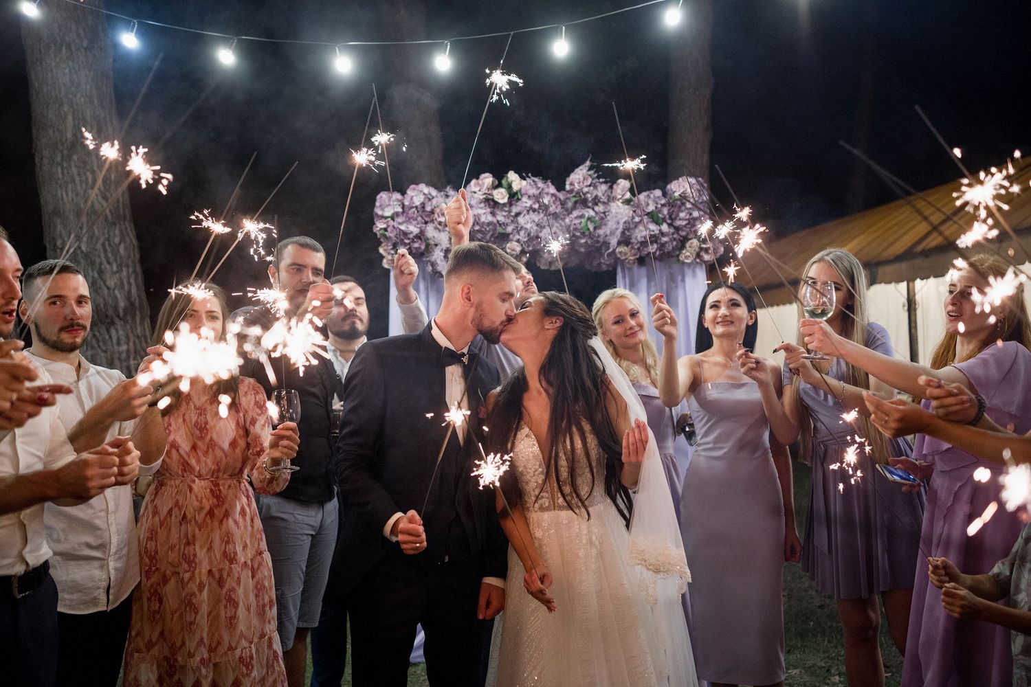 Bride and groom kissing surrounded by wedding guests holding sparklers at an outdoor night celebration.