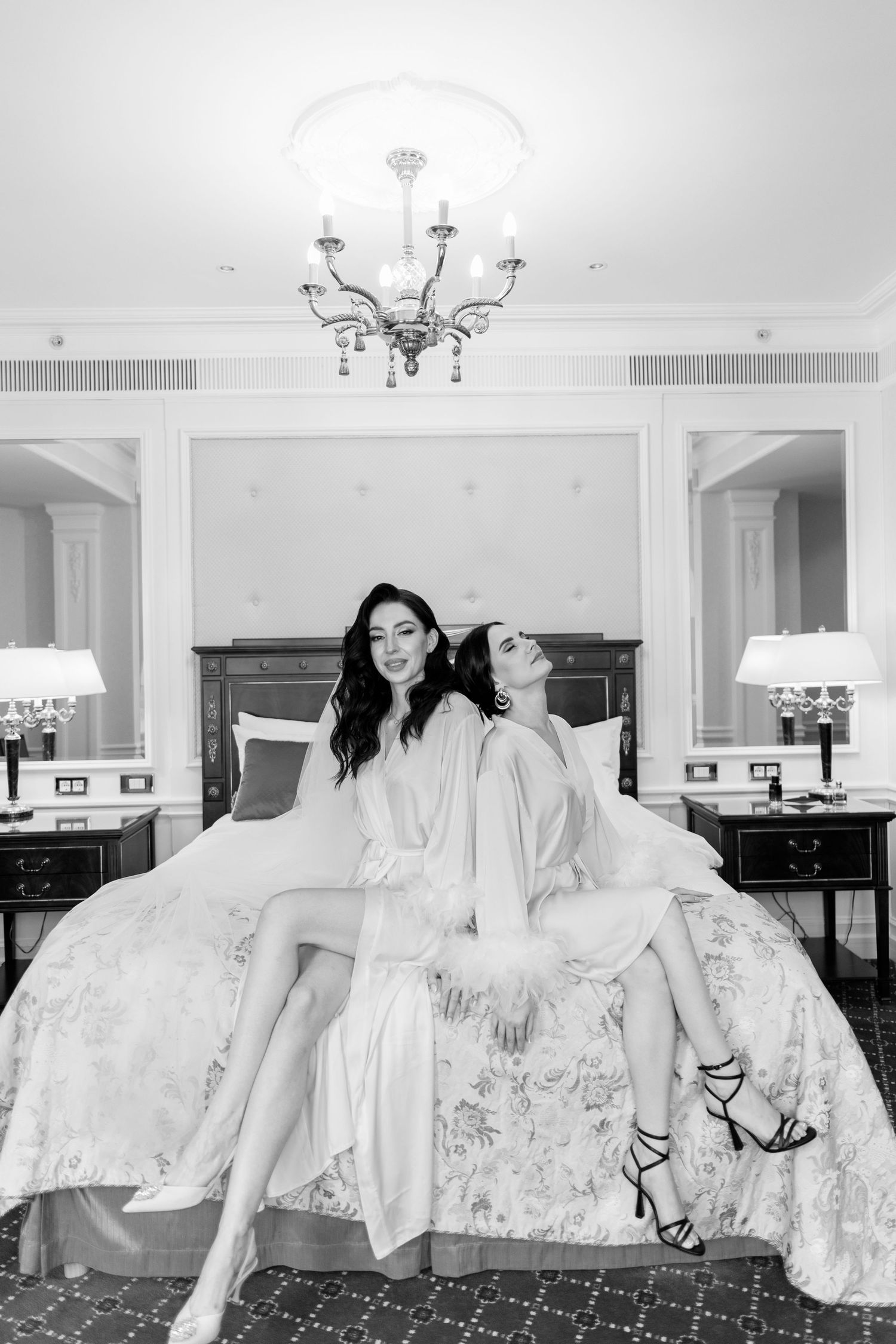 Two women in white robes sitting back-to-back on a bed in an elegant hotel room, black and white photo.
