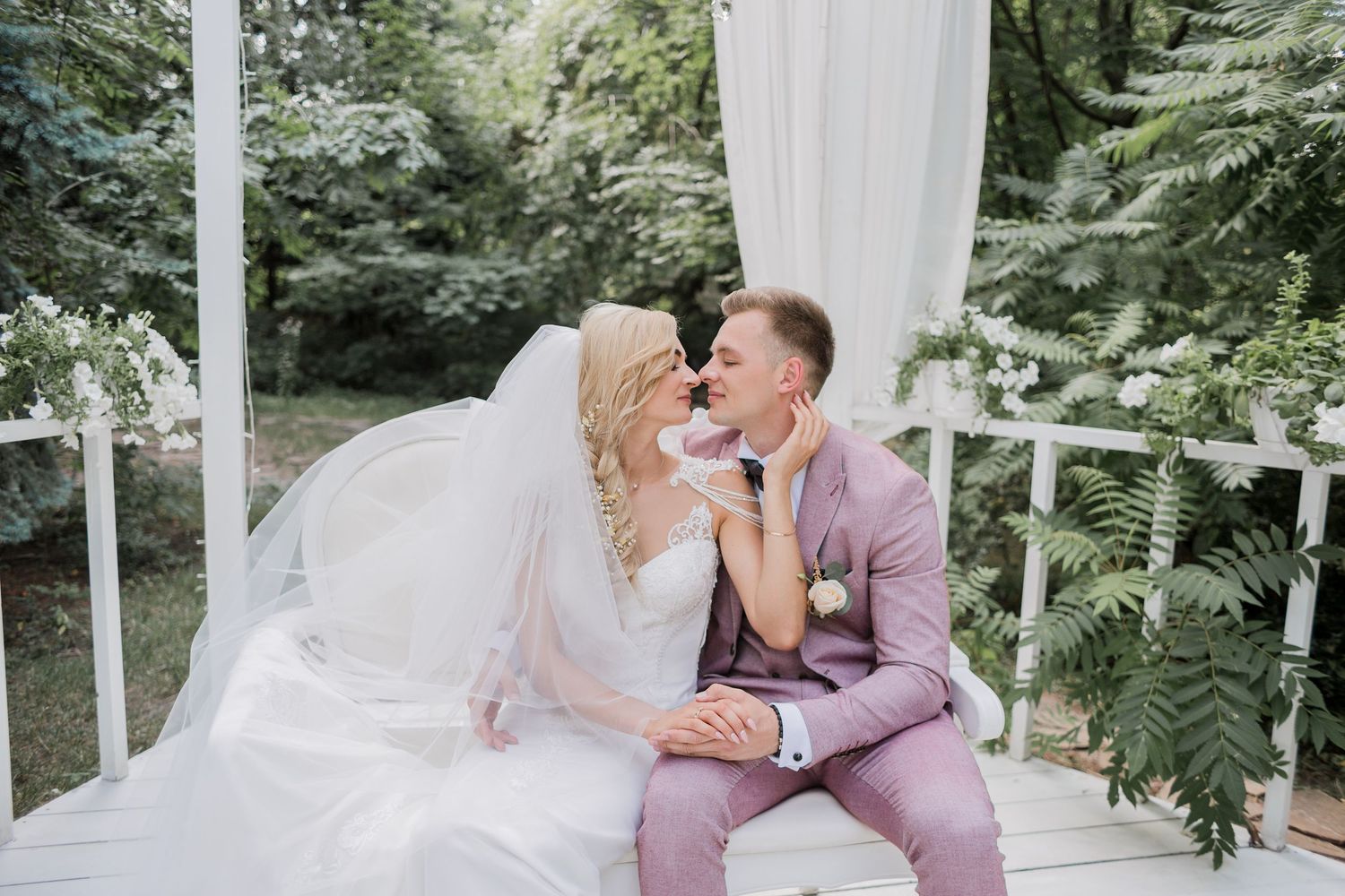A bride and groom share a romantic moment on a white gazebo, surrounded by lush greenery on their wedding day.