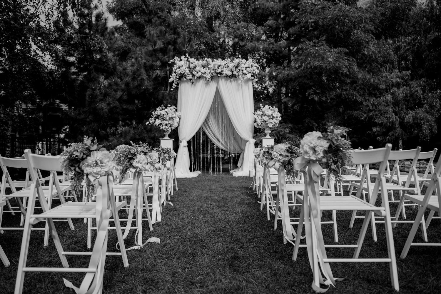 Black and white outdoor wedding ceremony setup with white chairs, floral arch, and draped fabric aisle surrounded by lush trees.