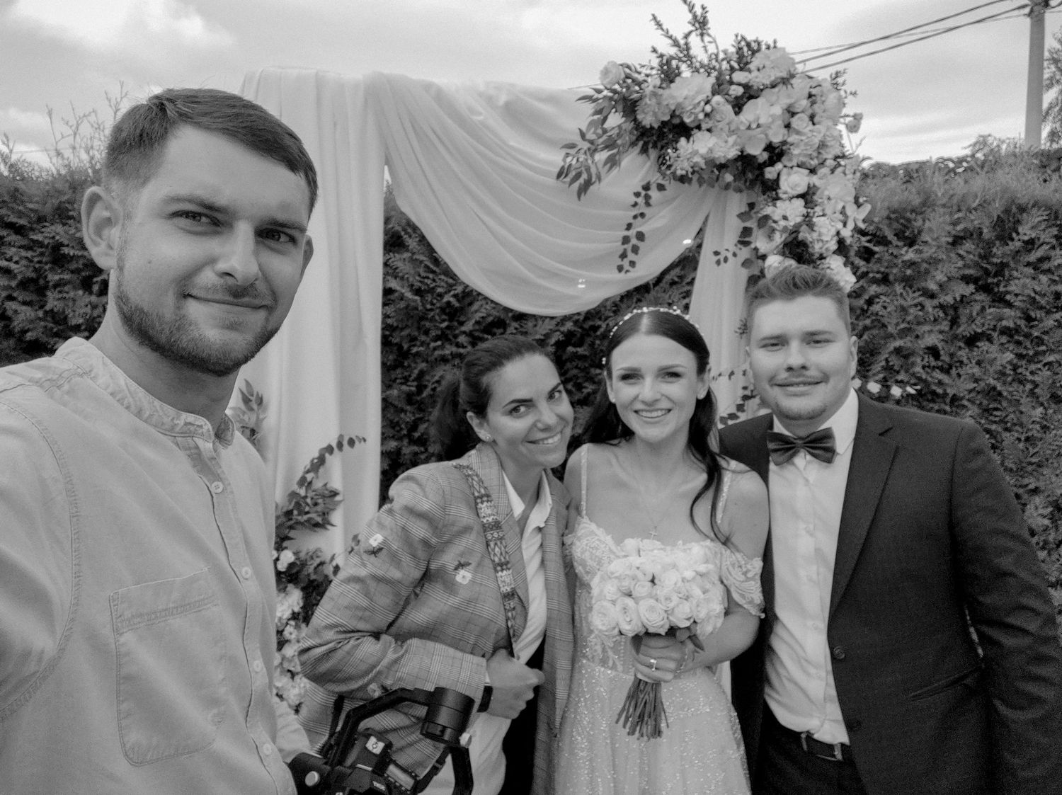 Black and white photo of four people posing together at an outdoor wedding ceremony with floral arch.