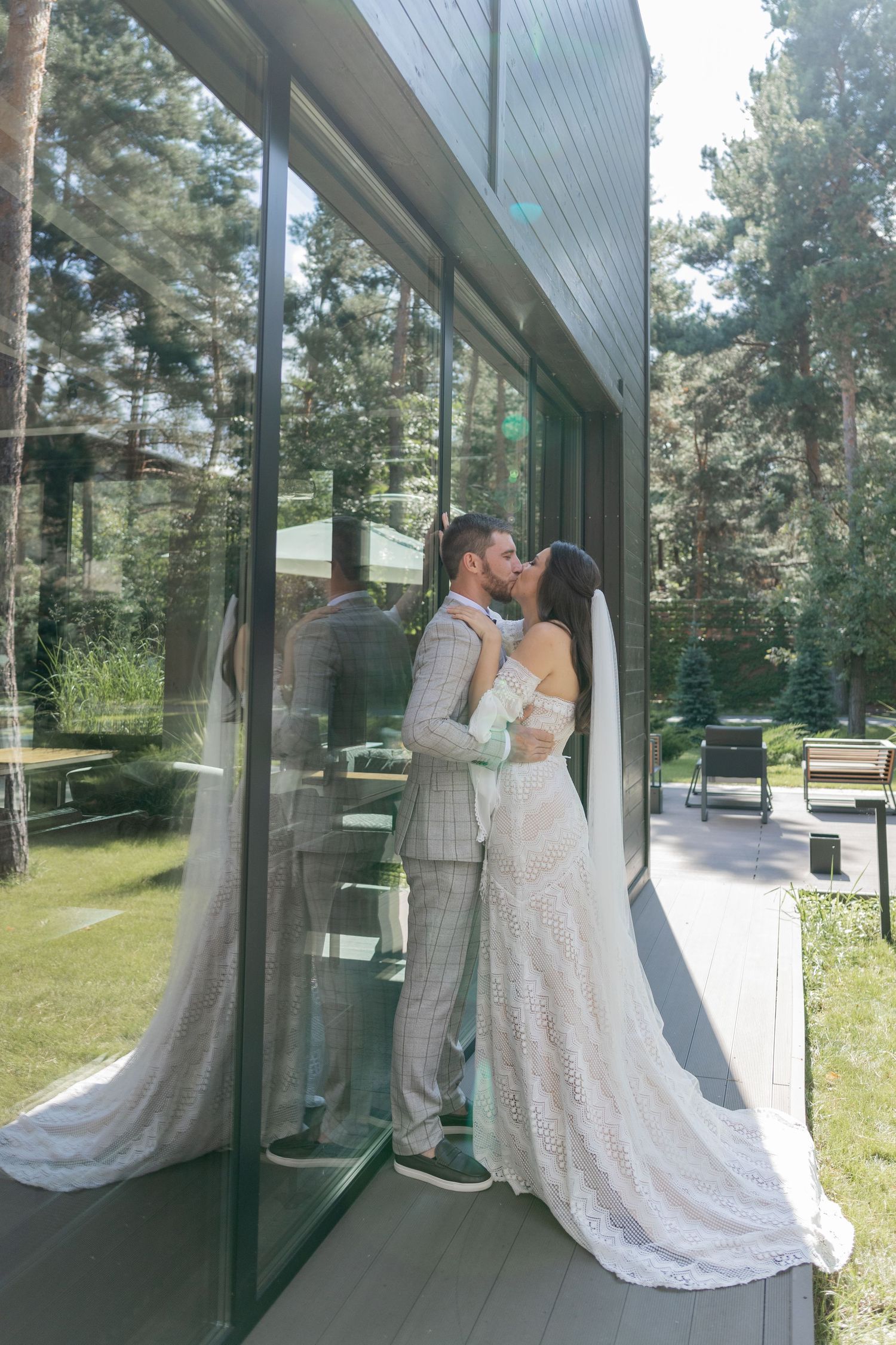 Wedding couple embraces near glass windows with forest reflections in black and white.
