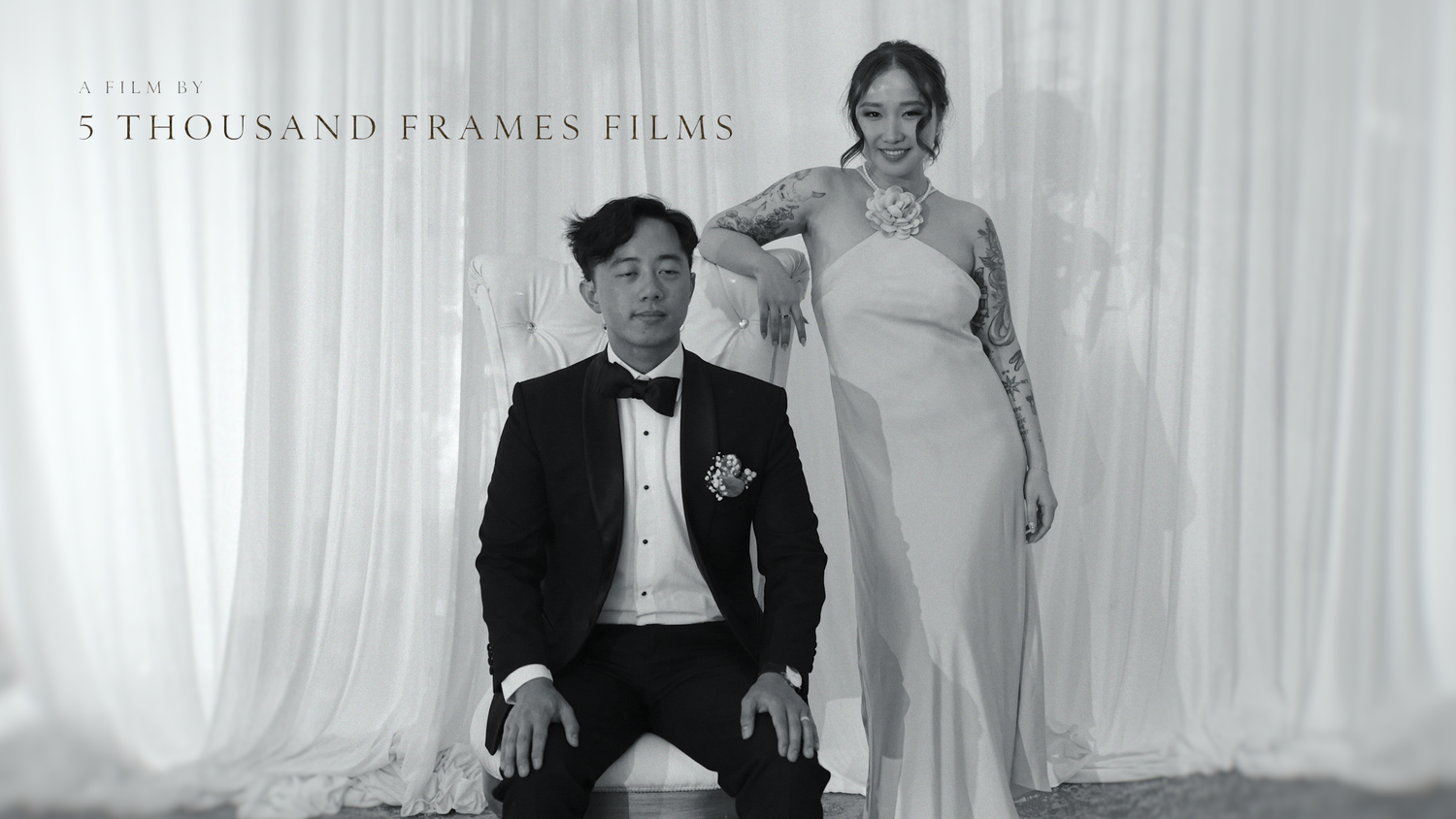 Black and white wedding photo of a couple, groom in tuxedo seated, bride in elegant gown standing, by 5 Thousand Frames Films.
