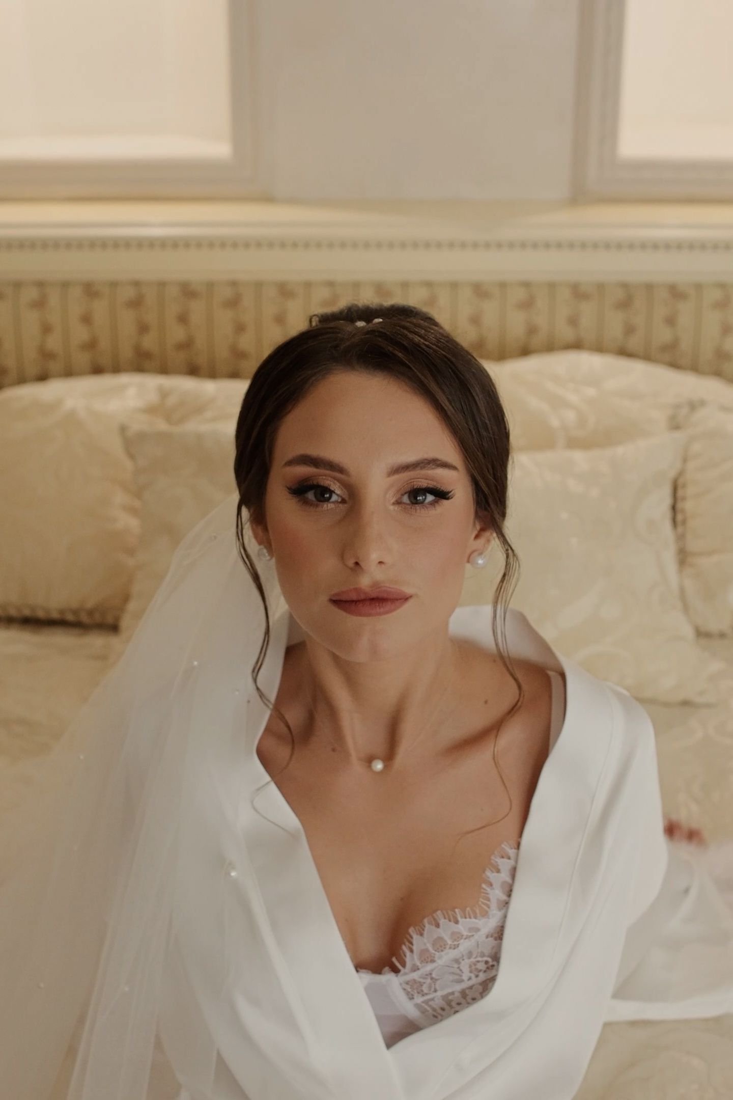 Bride in white robe and veil with elegant updo and pearl necklace sits gracefully before a luxurious cream bed.