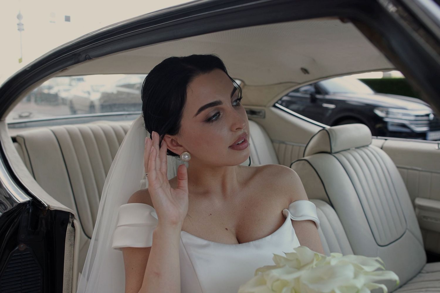 Elegant bride in white off-shoulder gown sitting in vintage car backseat, holding bouquet and adjusting earring.