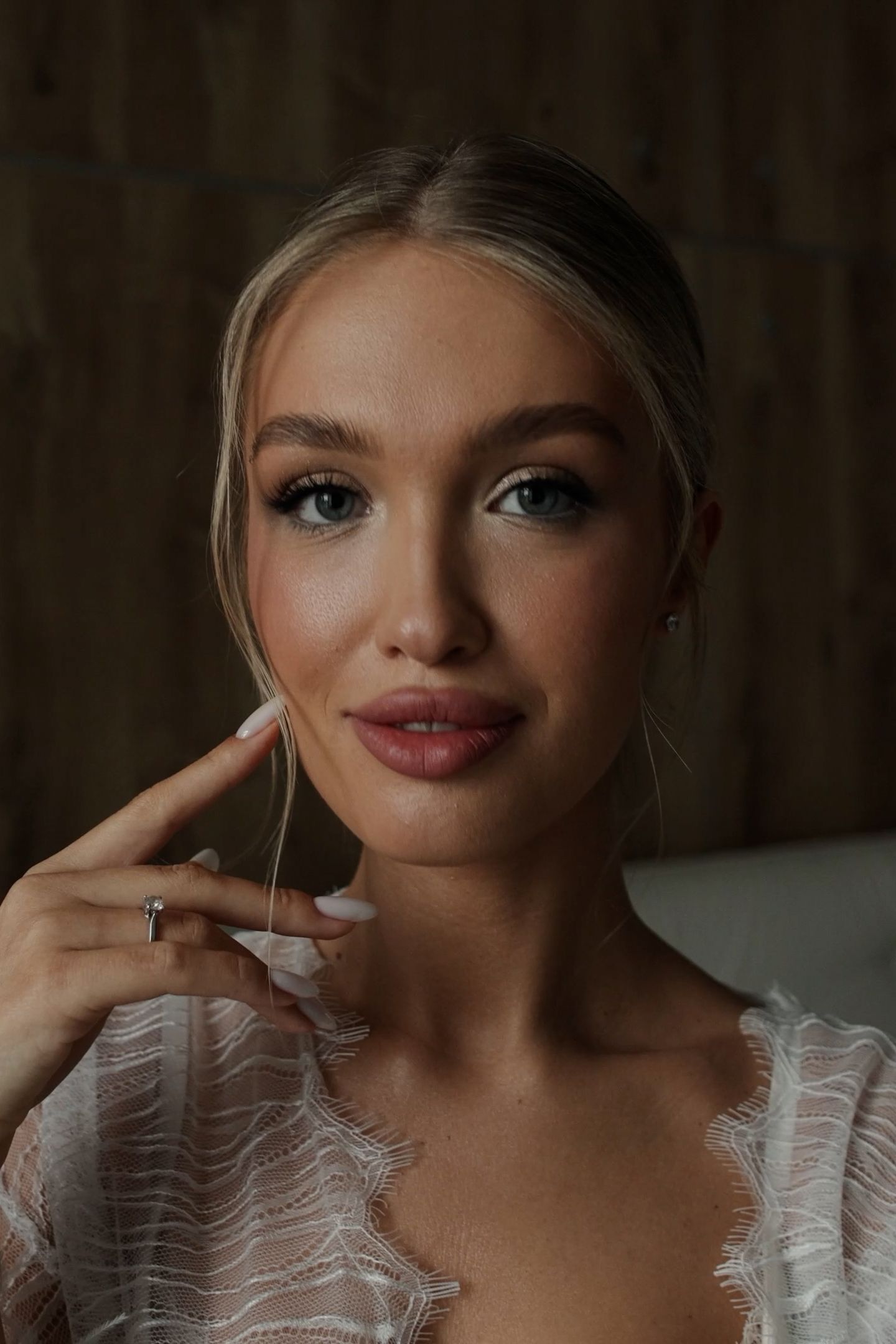 Young blonde woman with elegant makeup and white lace top poses gracefully touching her face with a ringed hand.