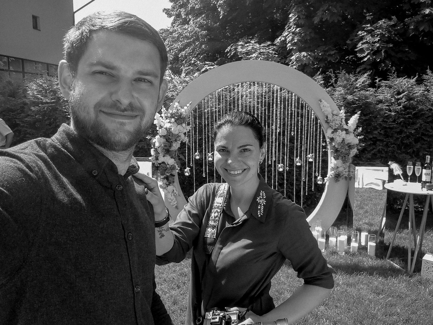 Black and white photo of a smiling man and woman posing outdoors near a decorative arch at an outdoor event.